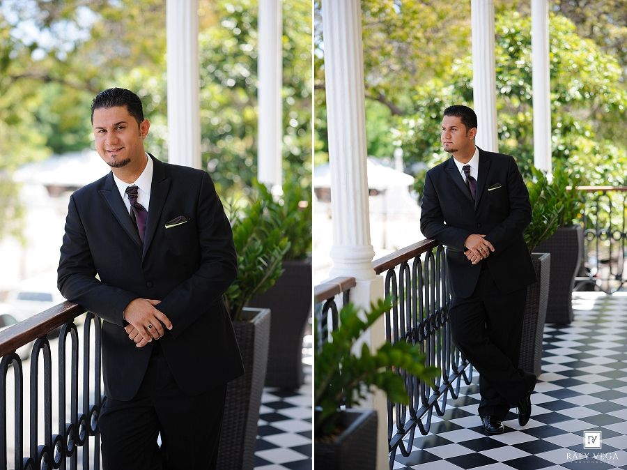 Dapper groom in black suit poses on elegant checkered porch with white columns and lush greenery.