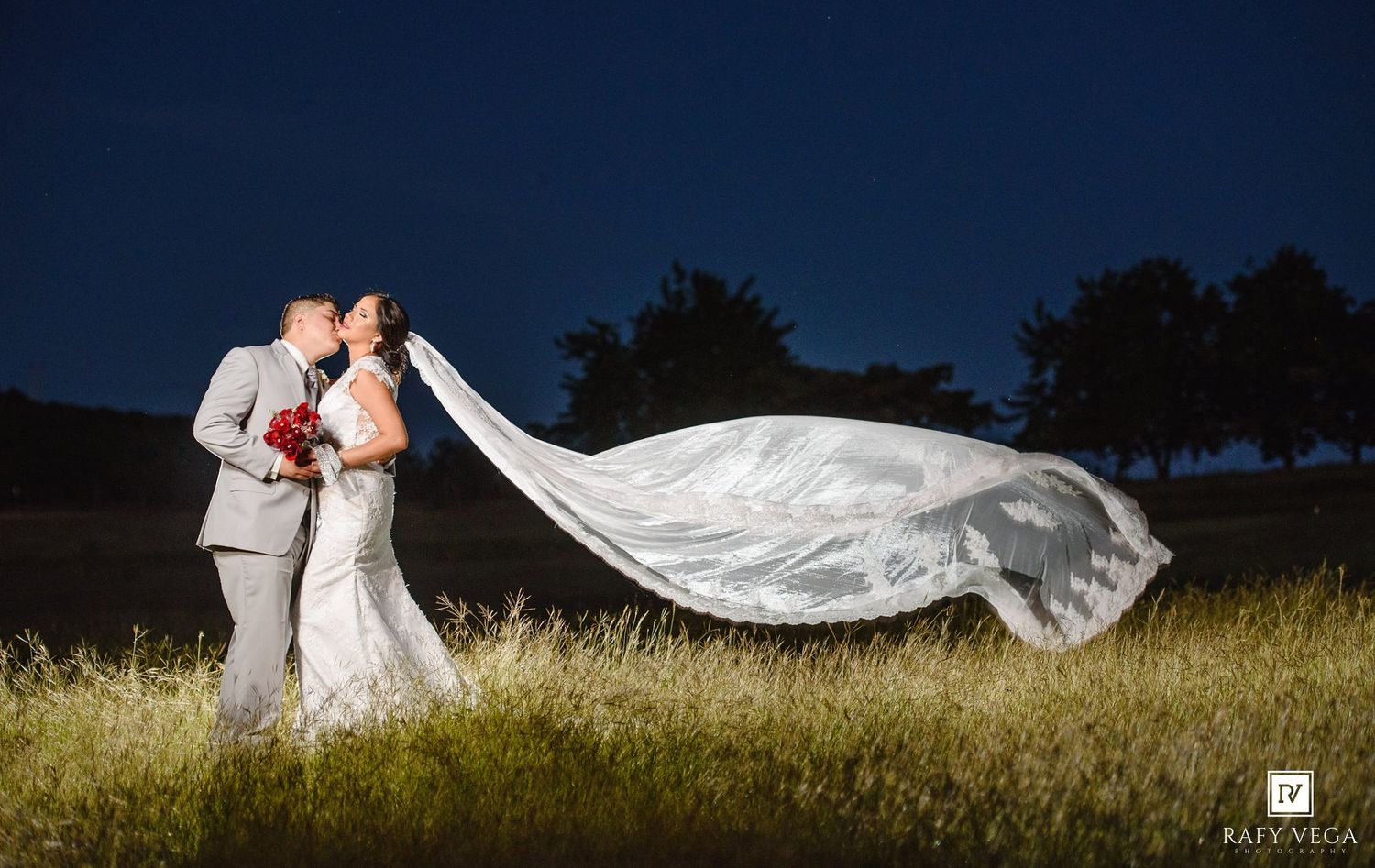 Bride and groom kissing at night with wedding veil dramatically flowing in the wind on grassy field.