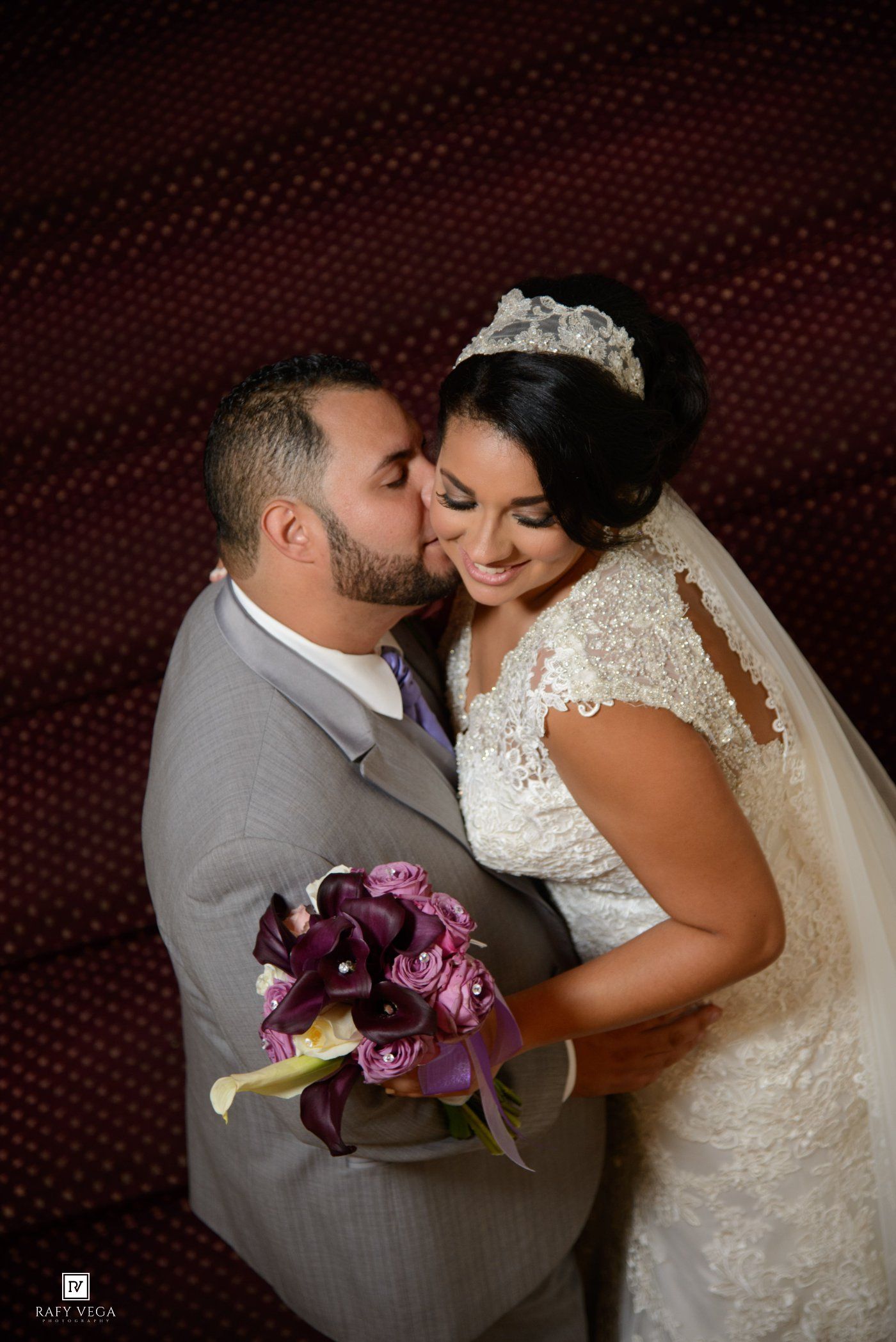 Bride and groom share a tender kiss, she holds purple calla lily bouquet, wearing lace gown and tiara.