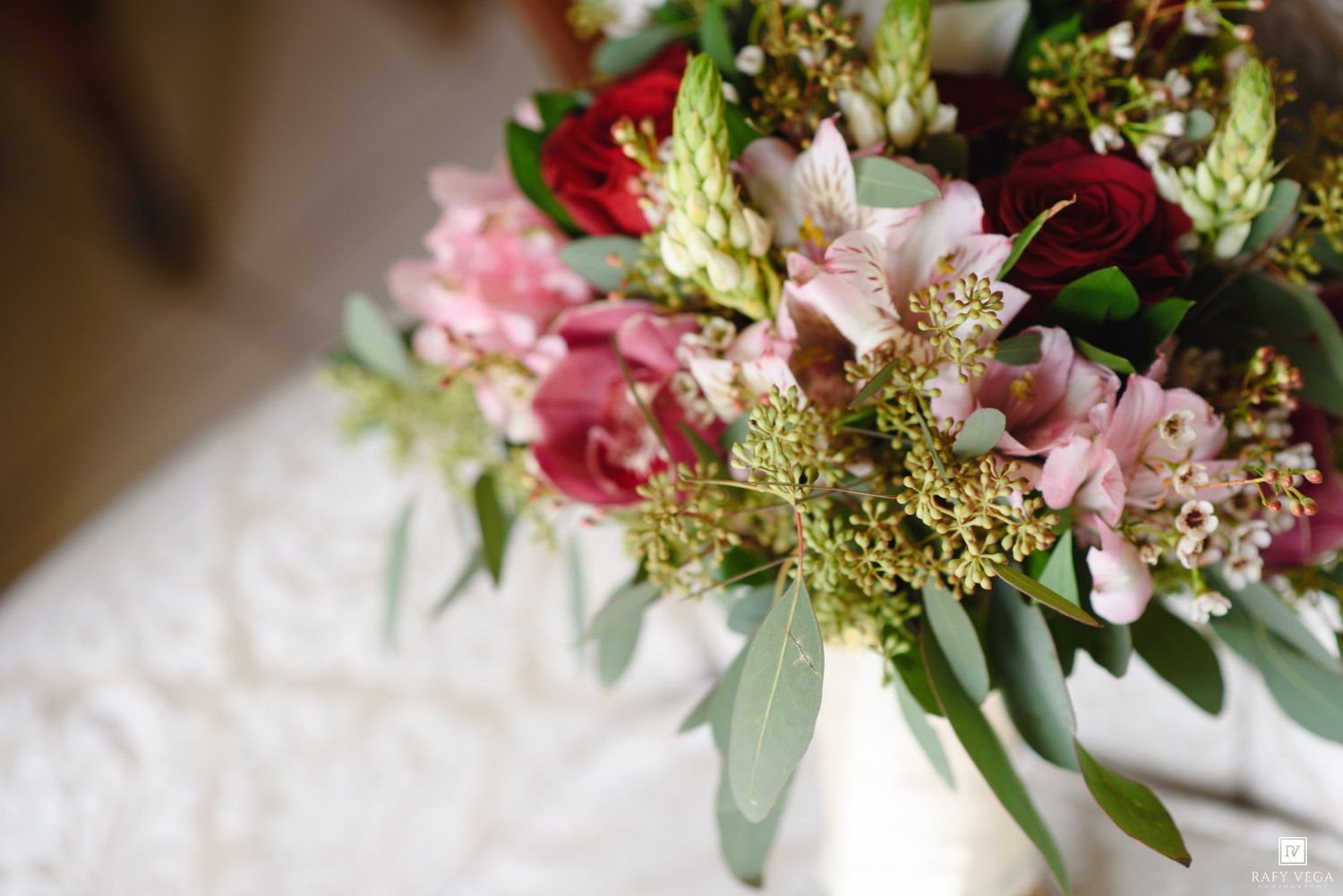 Close-up of a bridal bouquet with red roses, pink alstroemeria, and greenery in a white vase.