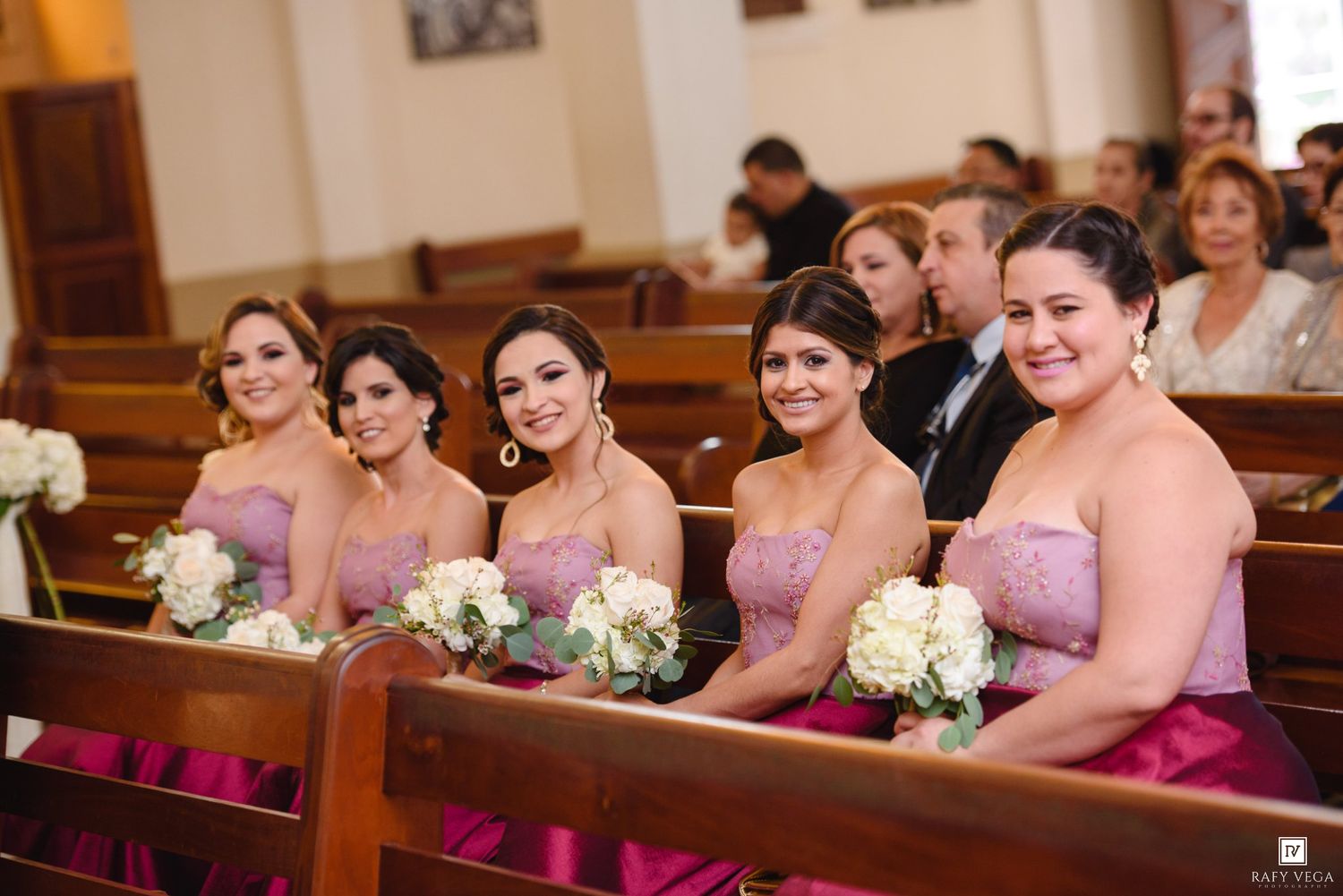 Five bridesmaids in pink strapless gowns holding white bouquets seated in church pews during a wedding ceremony.