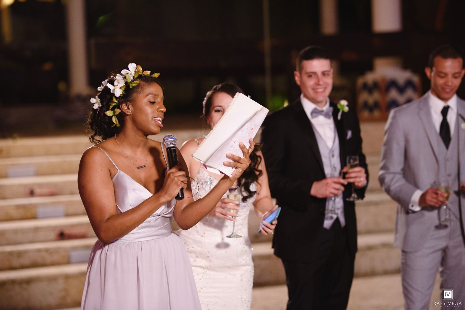Bridesmaid in floral crown gives speech at outdoor wedding reception while bride and guests listen.