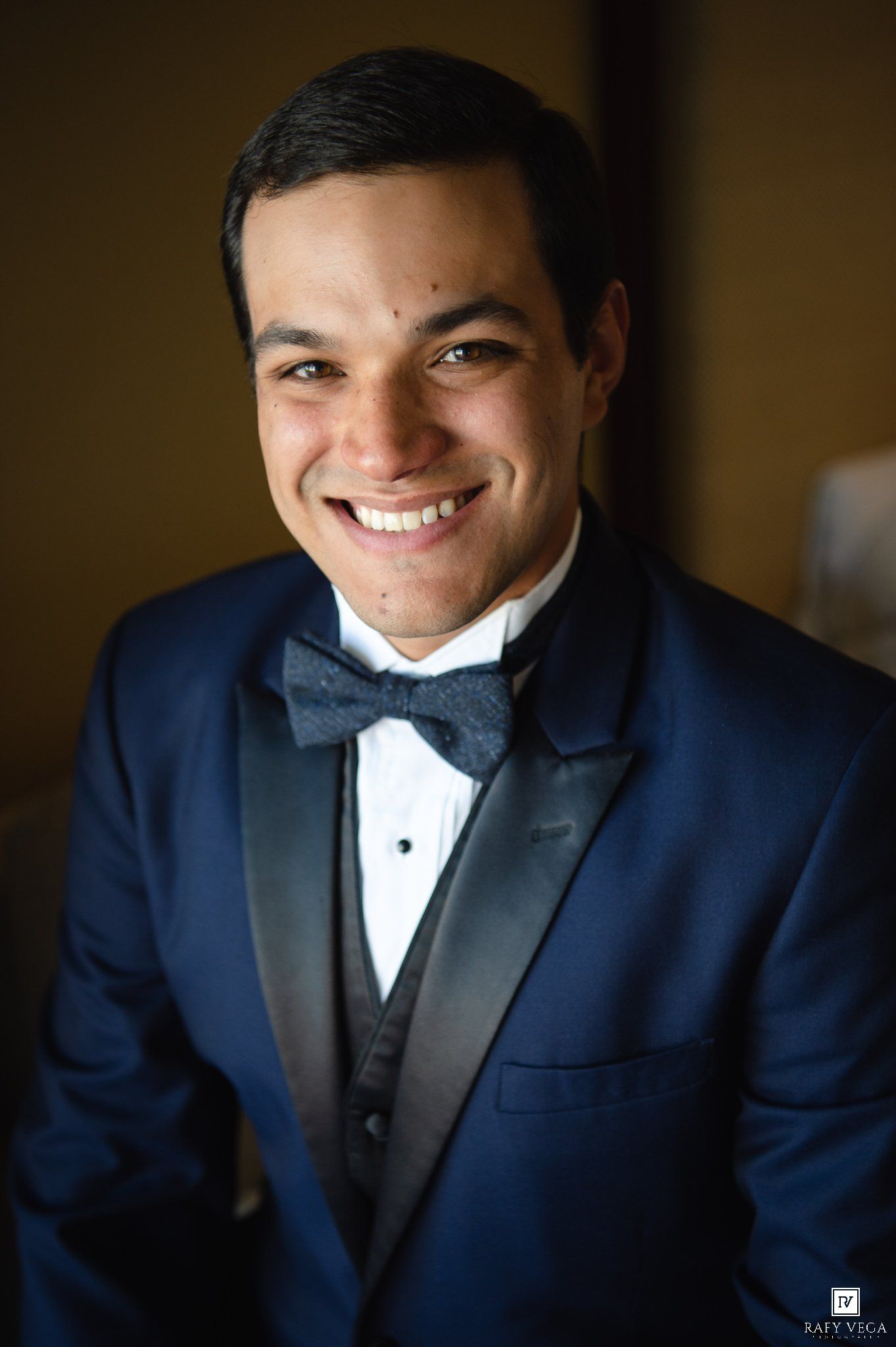 Smiling young man in navy blue tuxedo with black bow tie and vest, formal wedding attire portrait.