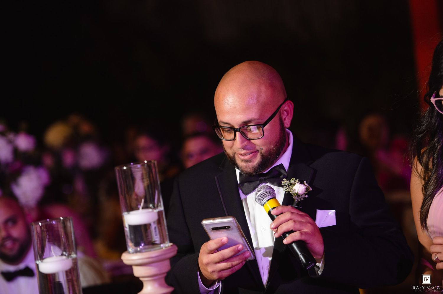 Bald man in tuxedo with boutonniere reads from smartphone while speaking into microphone at formal wedding reception.