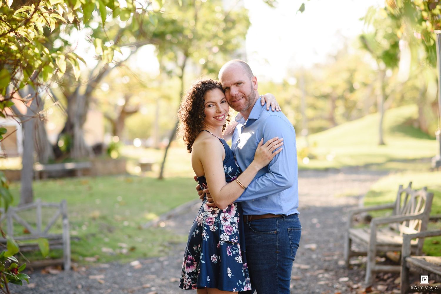 Couple embracing and smiling outdoors in a sunny park setting, with trees and wooden chairs in the background.
