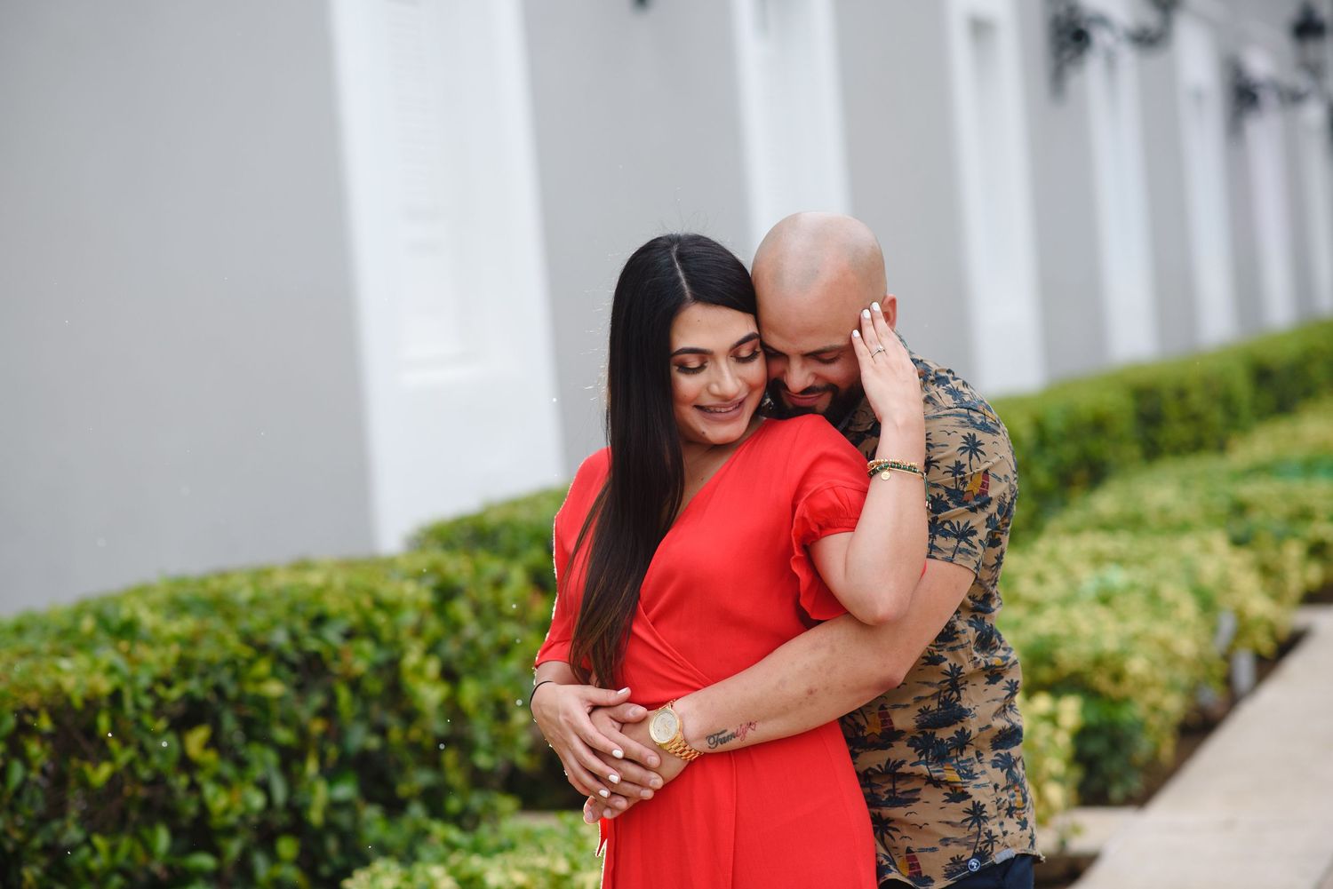 A couple embraces outdoors, woman in red dress with long dark hair, man in camo shirt, near green hedges.