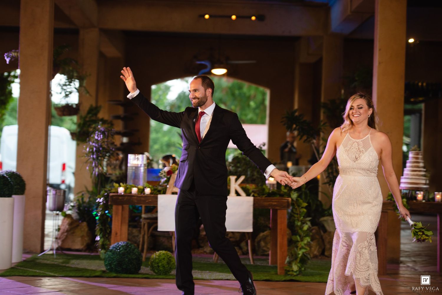 Bride and groom make grand entrance at outdoor wedding reception, groom waving to guests while holding bride's hand.