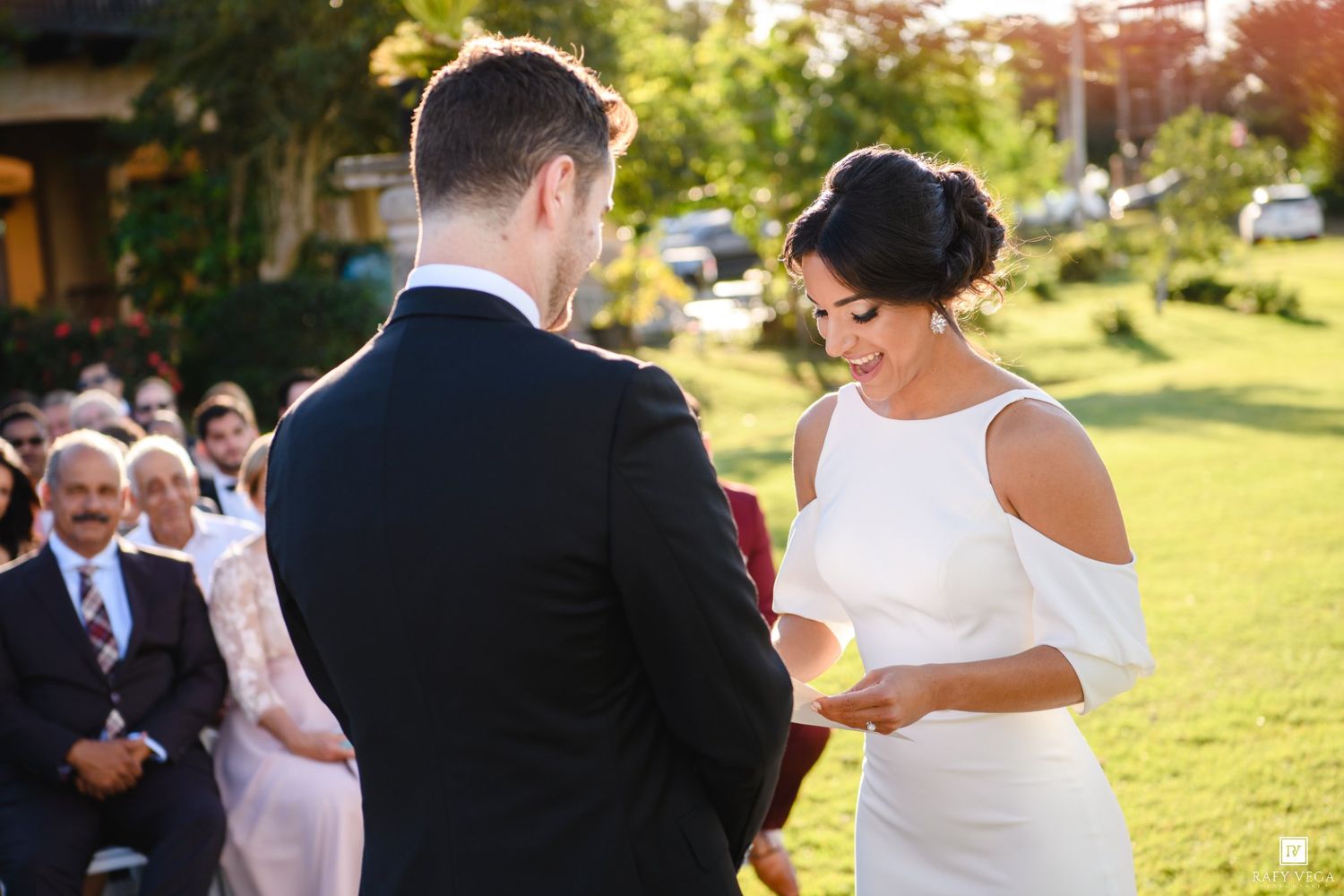 Bride reads wedding vows to groom during outdoor ceremony on green lawn with seated guests in background.
