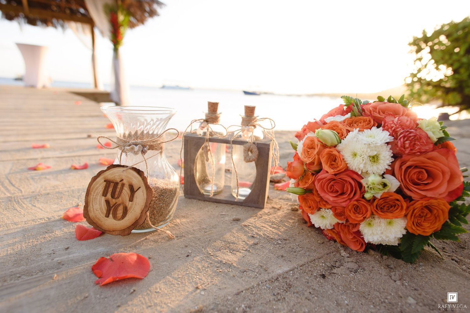 Tropical beach wedding decor with orange roses bouquet, sand ceremony bottles, and wooden TU YO sign on a wooden pier.