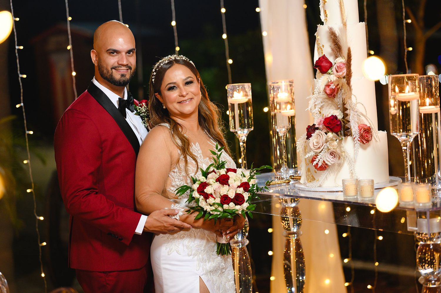 Newlyweds pose together by wedding cake with champagne flutes and fairy lights at night reception.