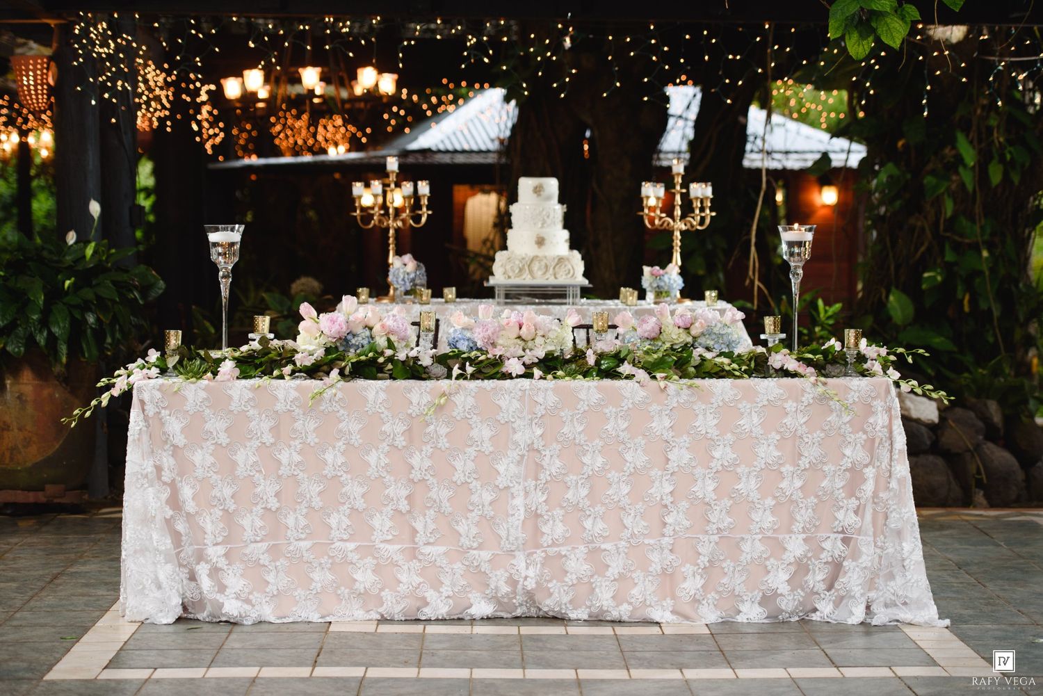 Elegant wedding reception table with white floral lace tablecloth, tiered cake, candelabras, and pink roses under twinkling lights.