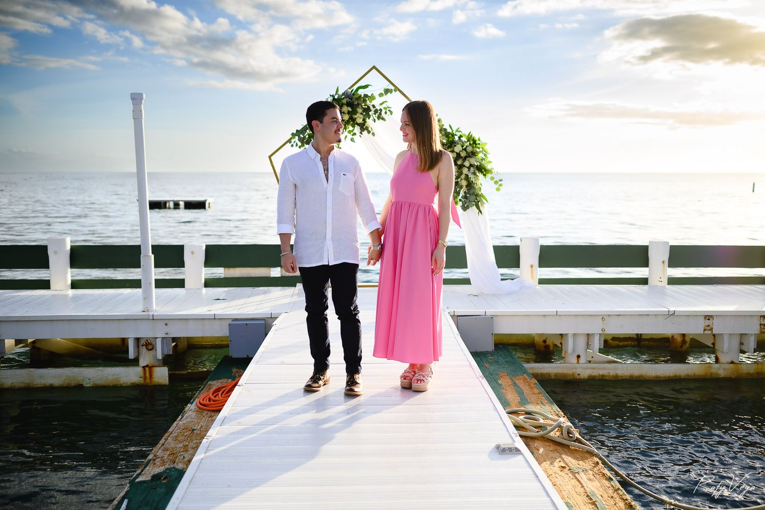 Couple walking hand in hand on a white pier dock with ocean backdrop and floral arch during golden hour.