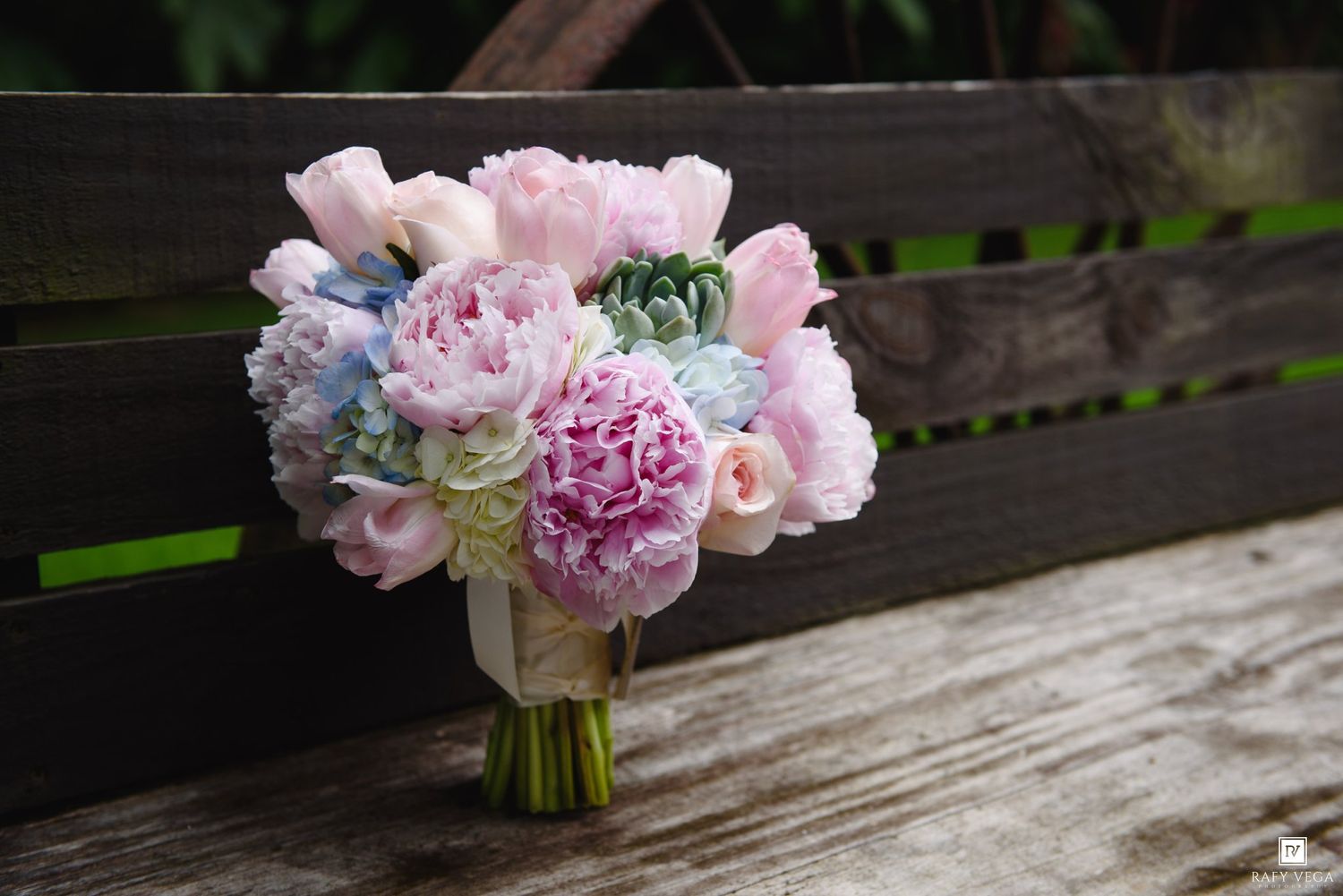 A romantic bridal bouquet of pink peonies, tulips, and blue hydrangeas resting on a rustic wooden bench.