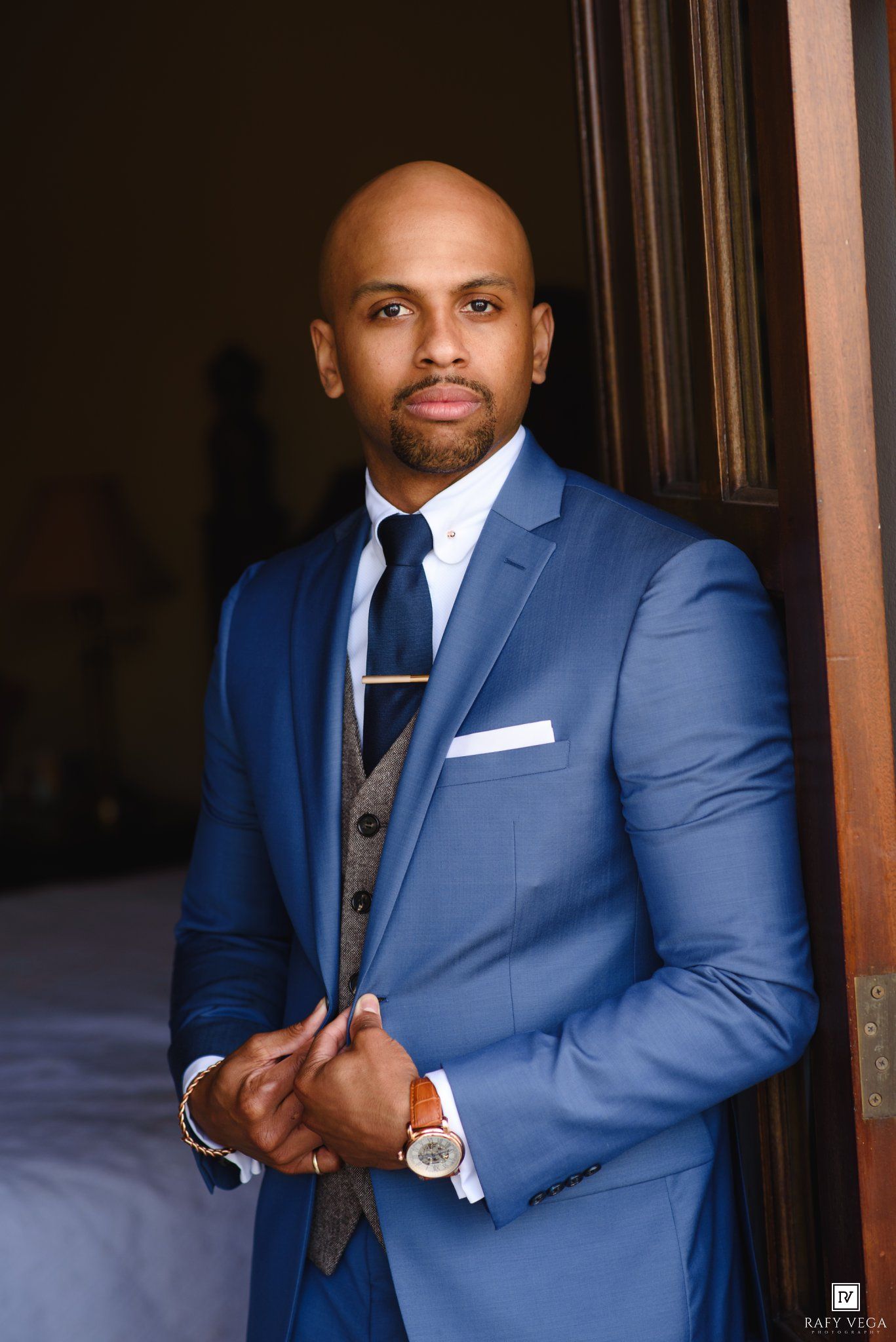 Elegant bald man in blue suit with gray vest, navy tie, and watch posing confidently in a doorway.