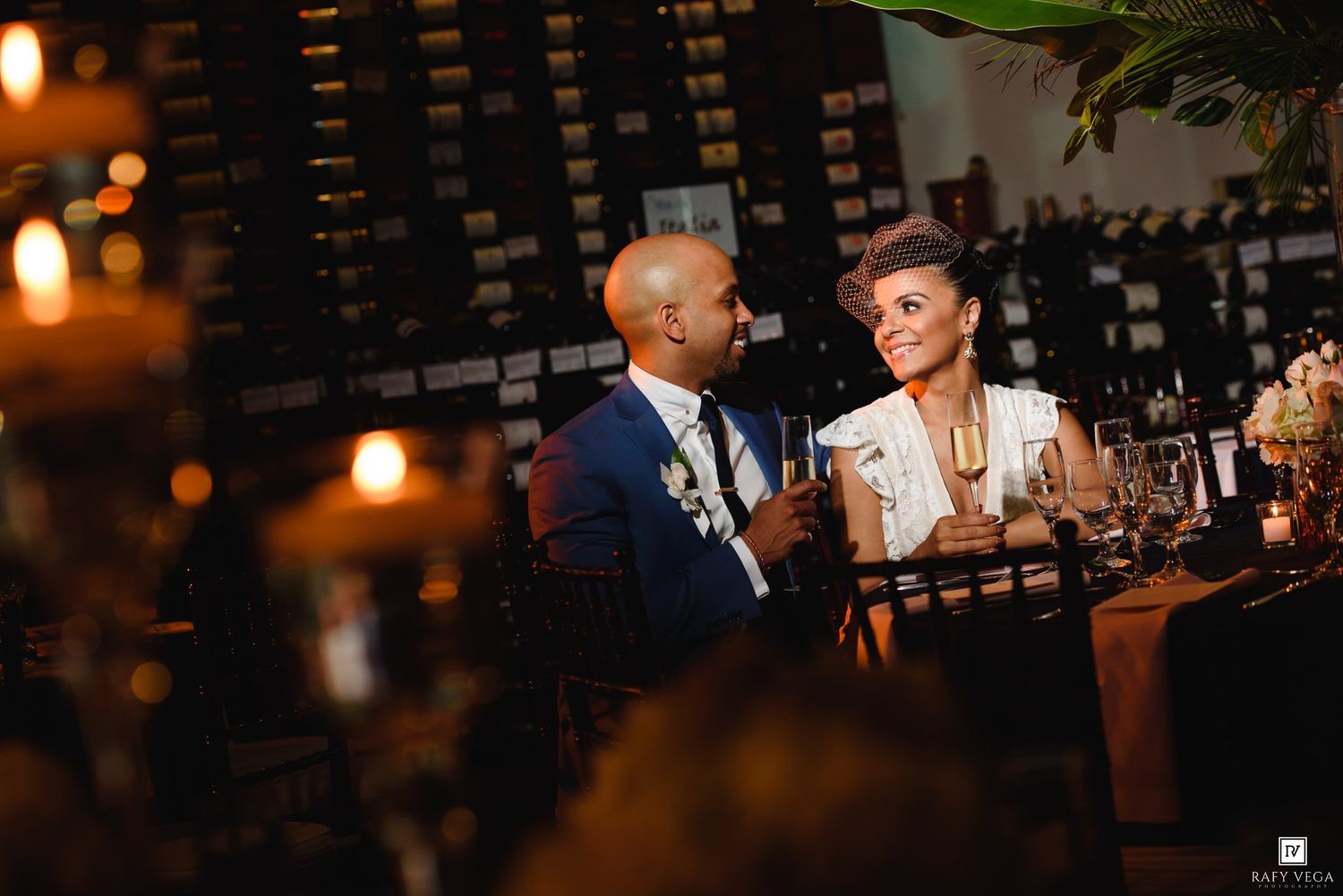 Couple in formal attire sharing a romantic moment at a candlelit wedding reception with wine wall backdrop.