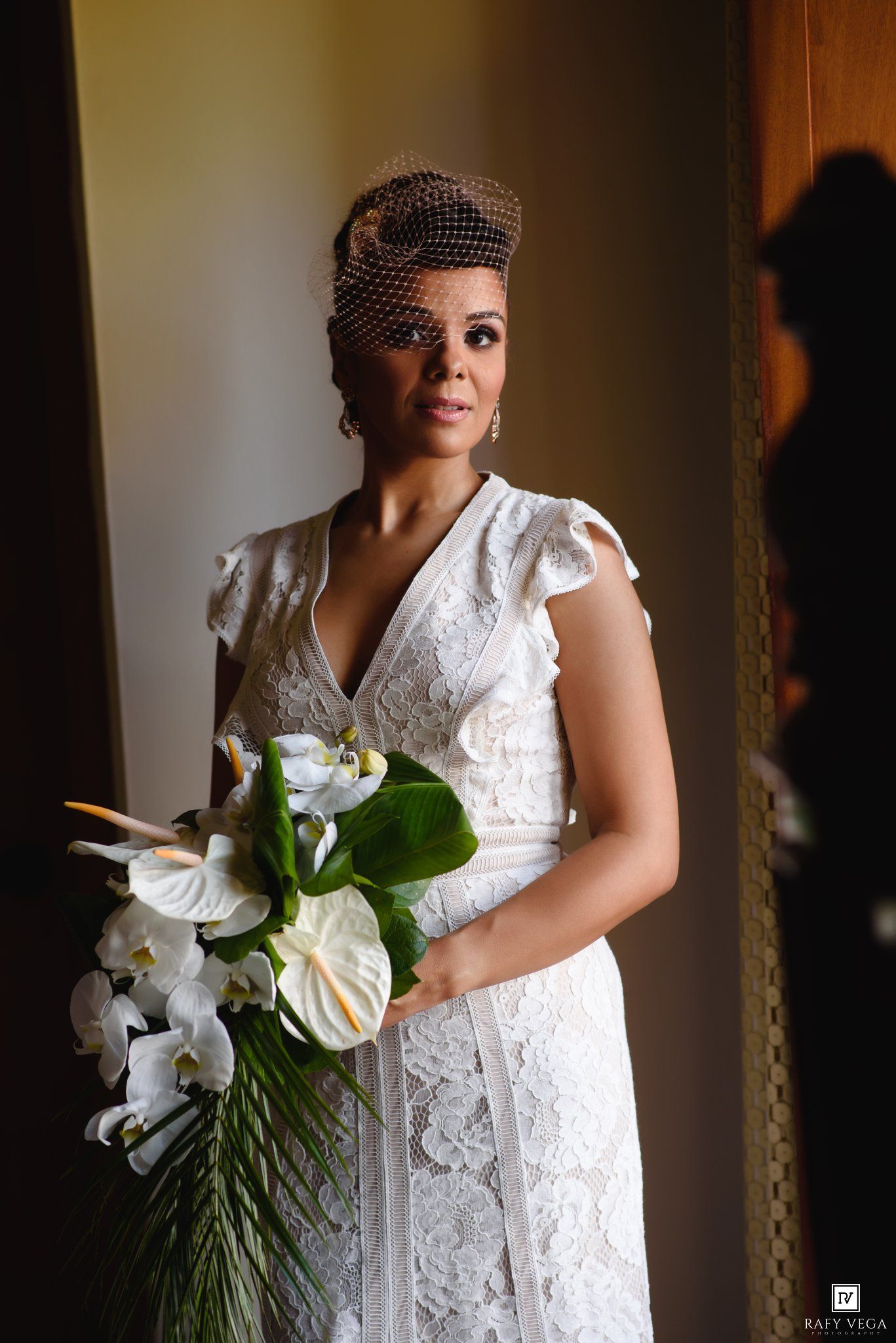 Elegant bride in white lace dress holding tropical bouquet with white orchids, wearing birdcage veil fascinator.