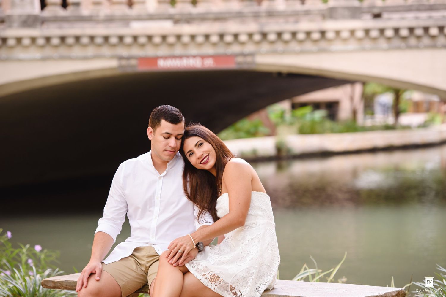 Couple sitting together by a canal with a stone bridge in the background on a sunny day.