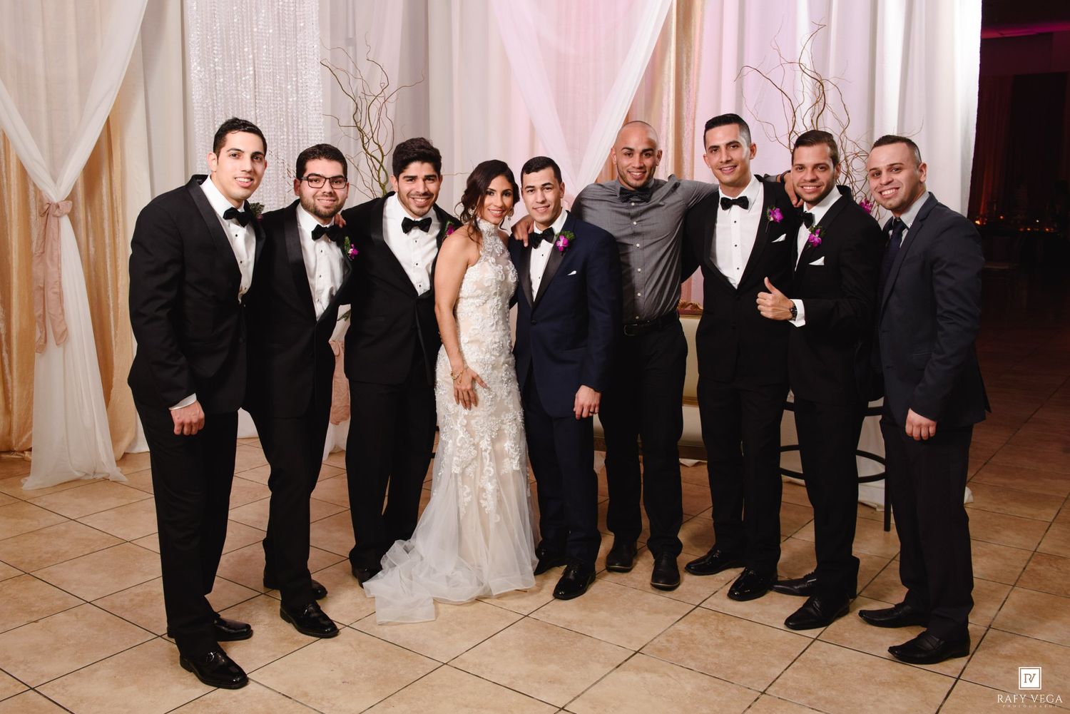 Wedding party poses together at elegant reception, bride in white gown with groomsmen in black tuxedos.