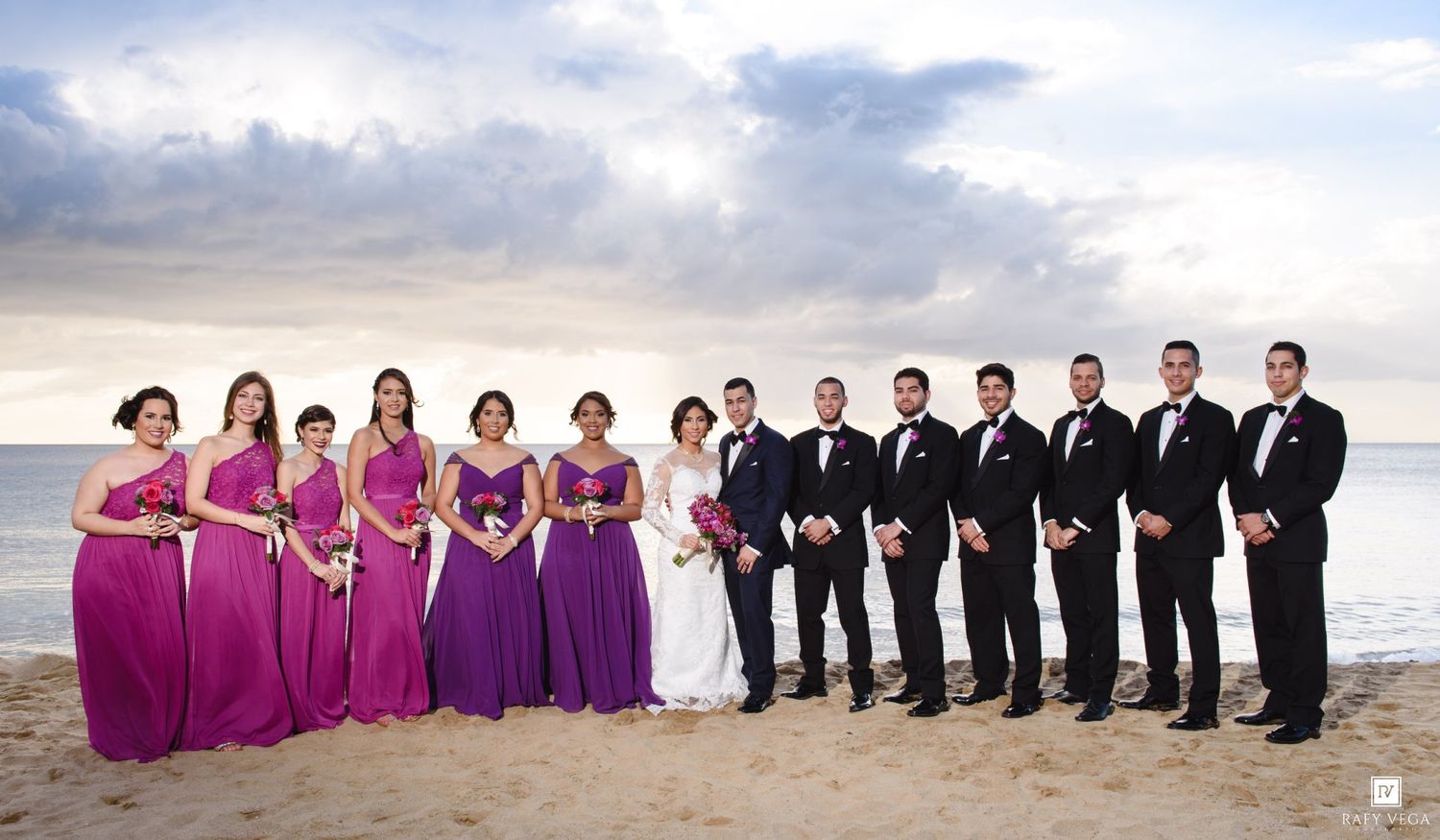 Wedding party poses on beach, bridesmaids in purple gowns and groomsmen in black tuxedos, with bride and groom centered.