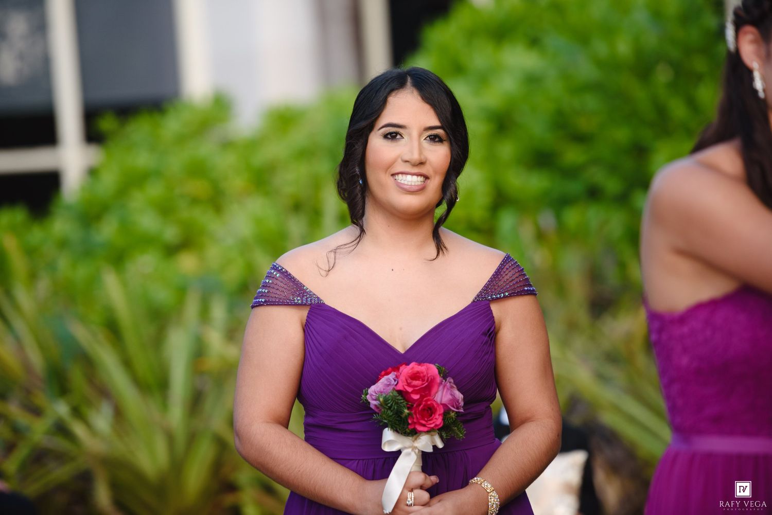 Smiling bridesmaid in purple off-shoulder gown holds pink rose bouquet at outdoor wedding.