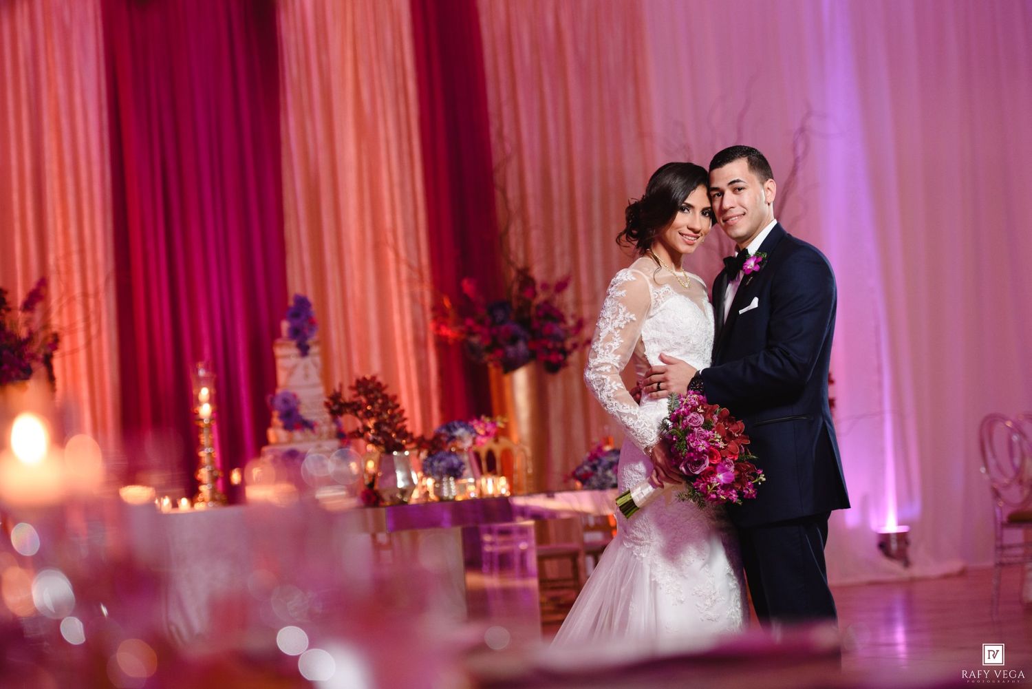 Bride and groom share romantic embrace at elegant wedding reception with pink draping and candlelit tables.