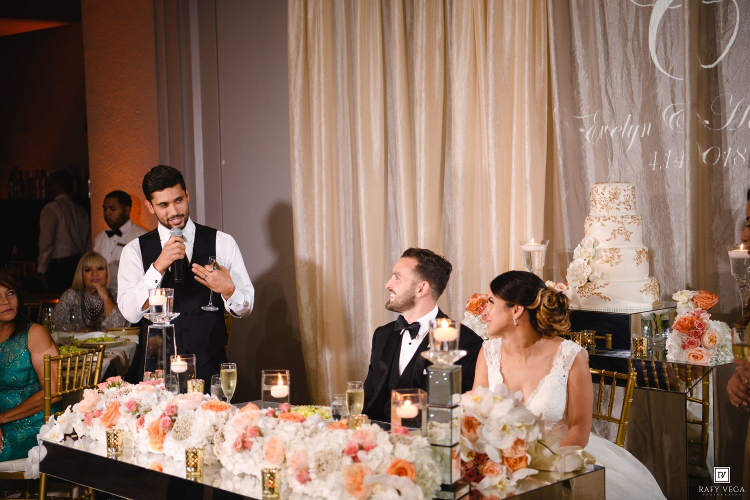 A groomsman delivers a toast at an elegant wedding reception with floral arrangements and a tiered cake.