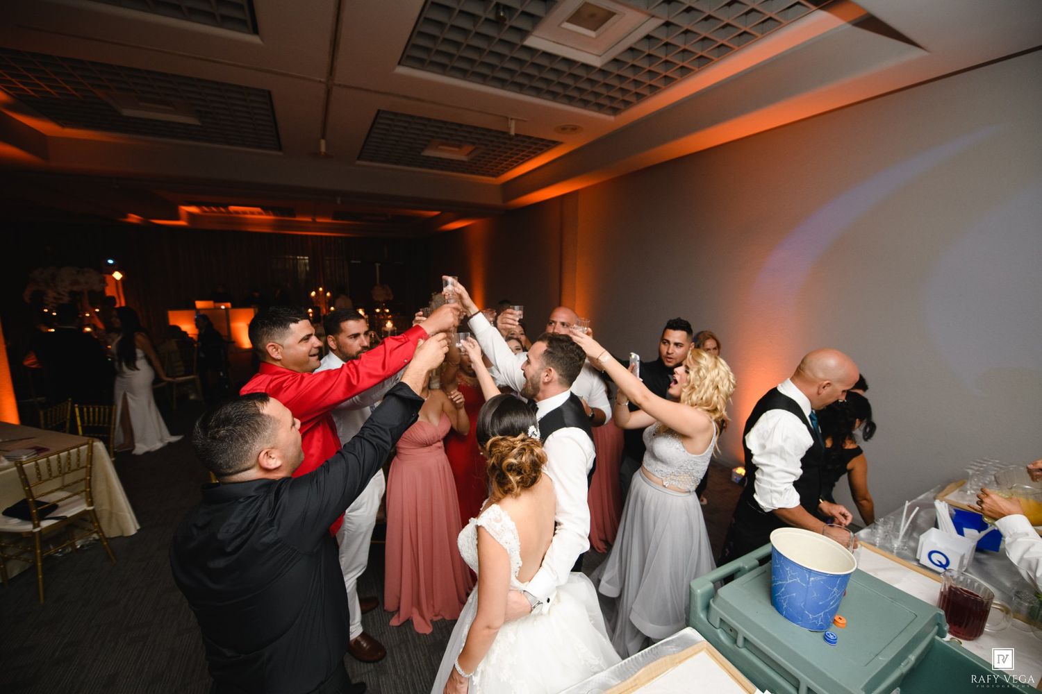 Wedding guests dance together in a circle on the reception dance floor under warm amber lighting.