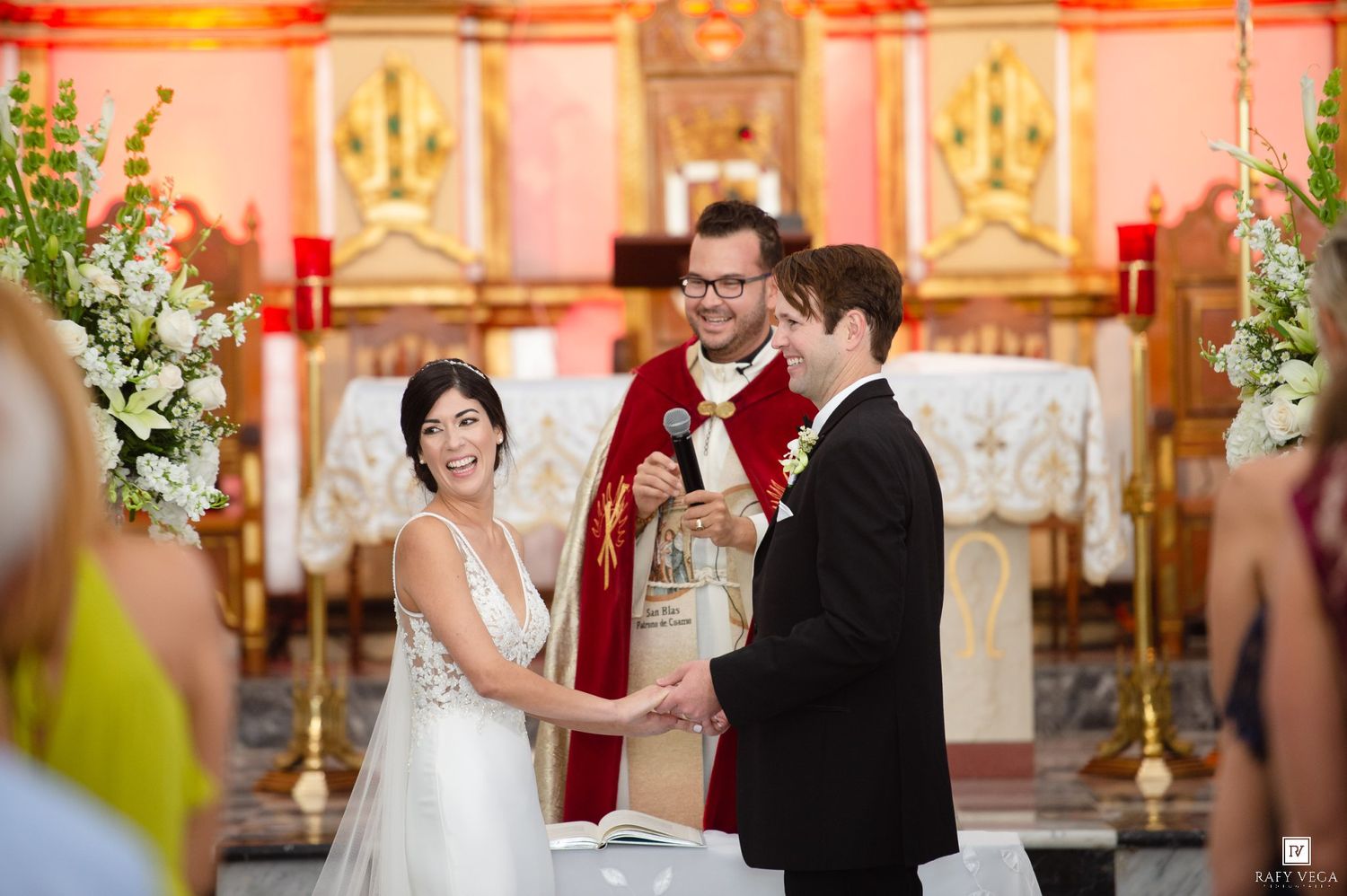Bride and groom exchange vows at a church altar with a priest in red vestments during a wedding ceremony.