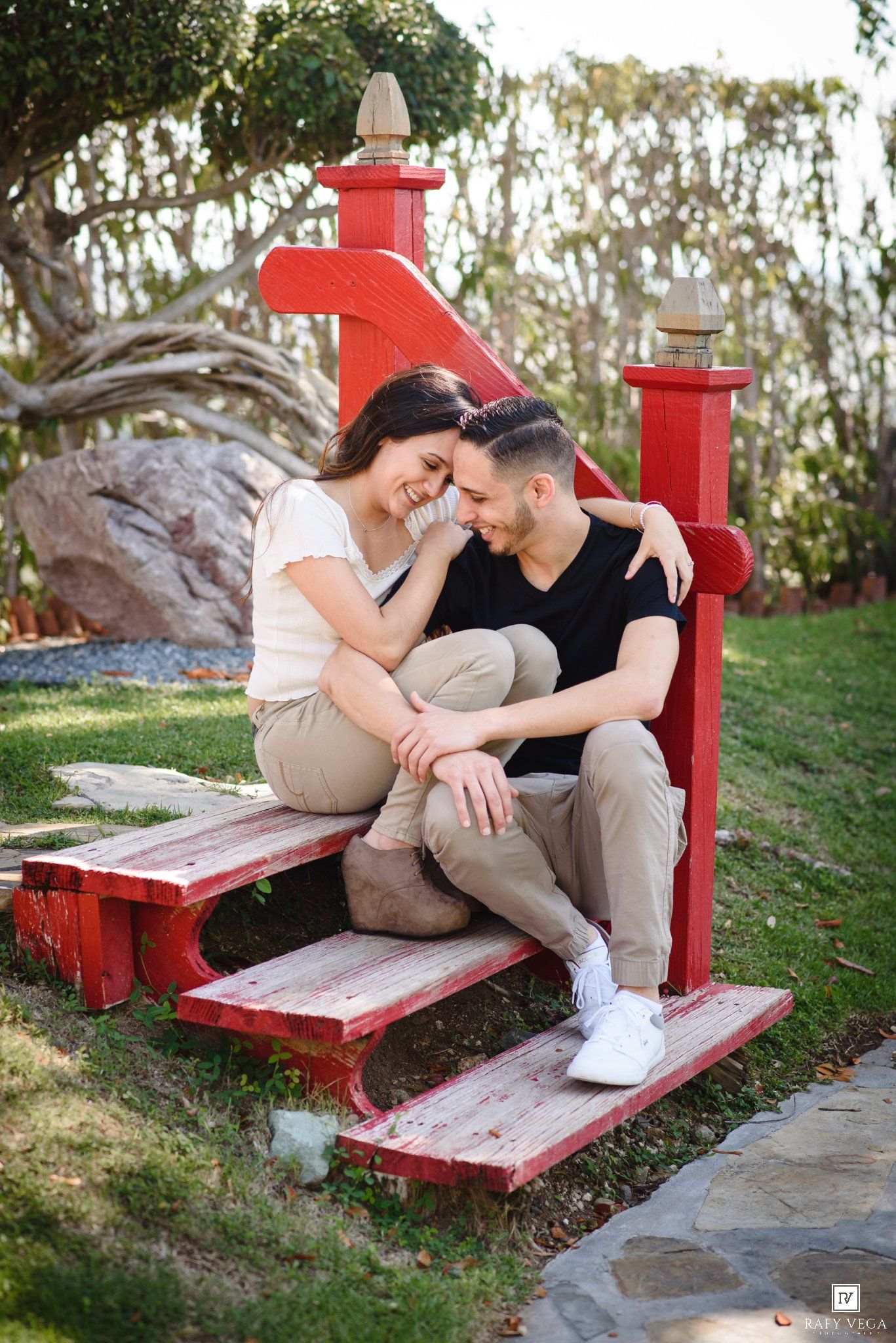 A happy couple sits closely together on a red wooden bench in a lush garden, smiling and embracing affectionately.