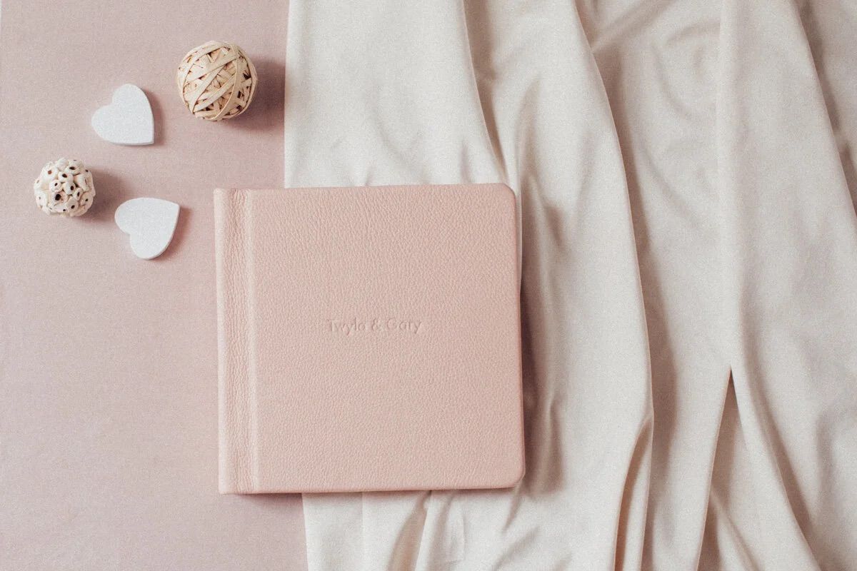 Pink linen photo album on white fabric with heart decorations and wicker balls on a blush background.