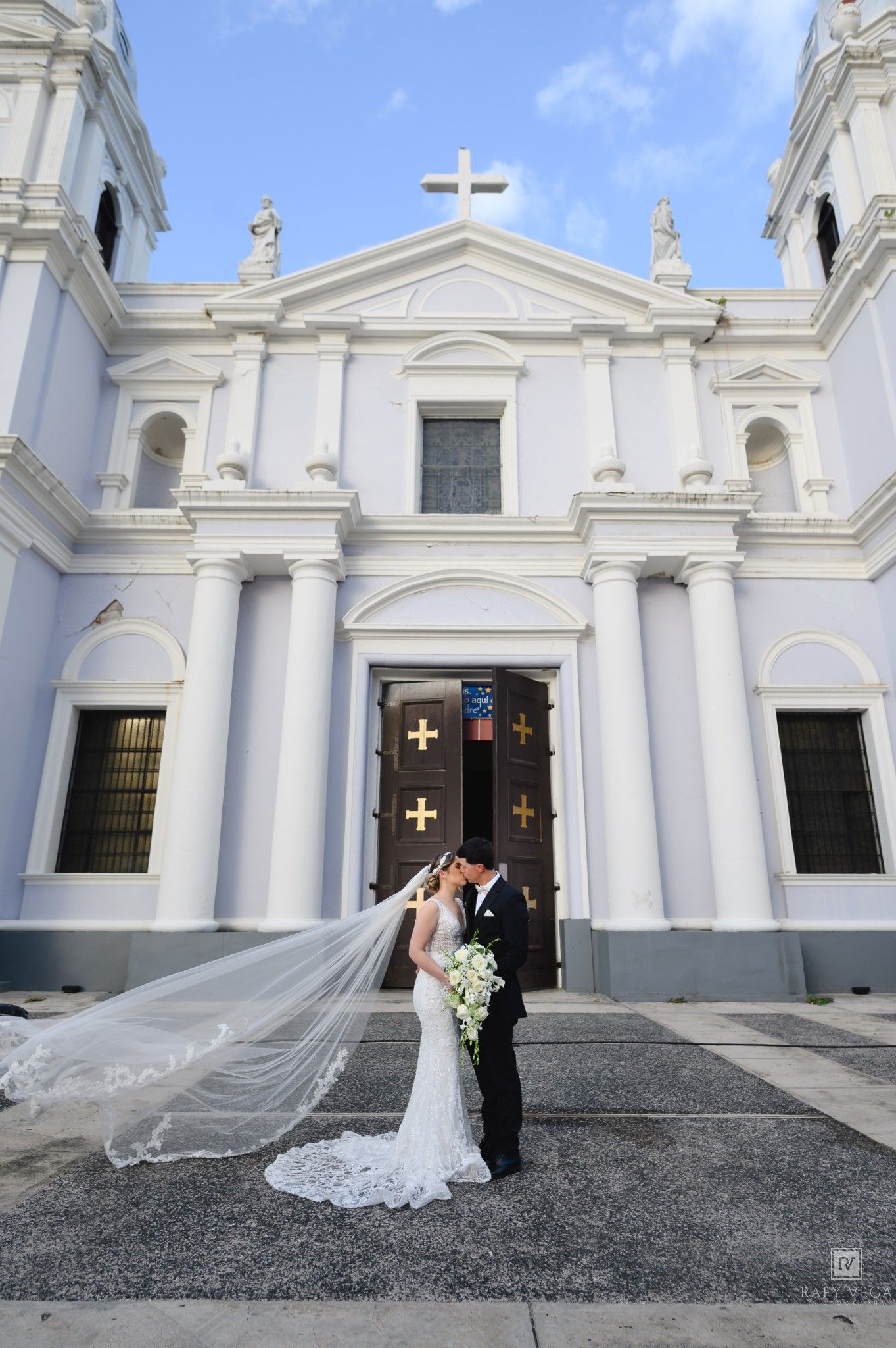 Bride and groom kissing in front of a white colonial church with golden cross doors, wedding veil flowing in the wind.