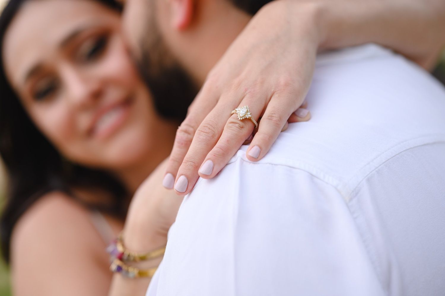 Woman showing off gold engagement ring with manicured nails while embracing man in white shirt outdoors.