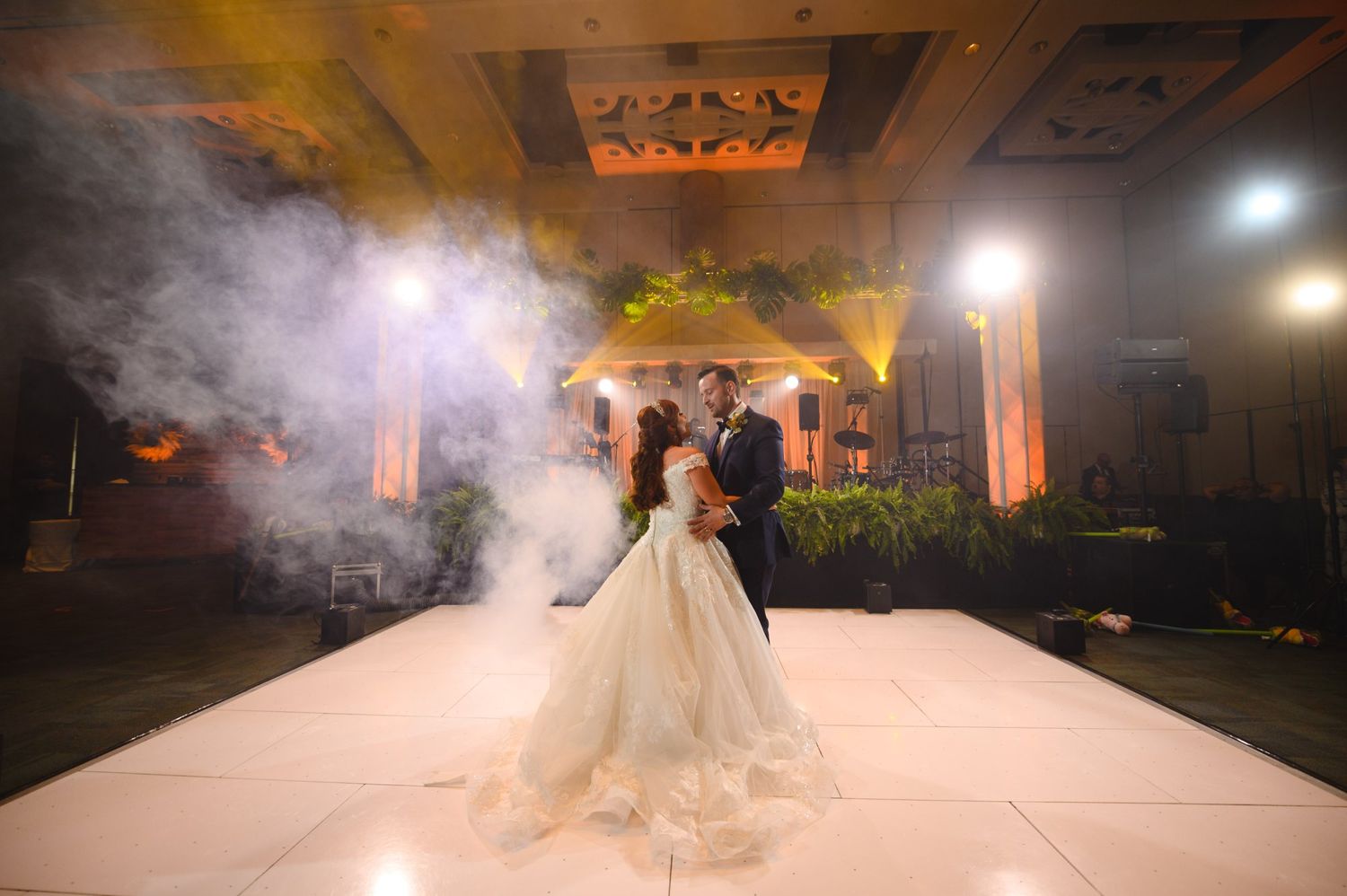 Bride and groom share first dance on white floor surrounded by dramatic fog effects at elegant wedding reception.