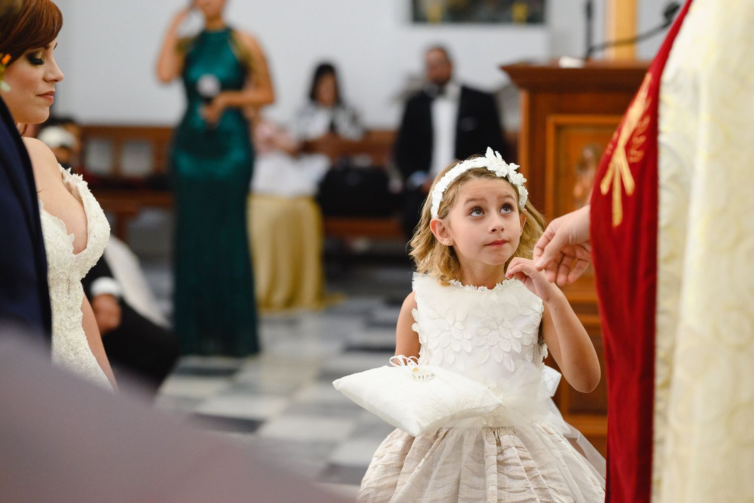 Young flower girl in white lace dress holding pillow at wedding ceremony, looking up with curiosity.