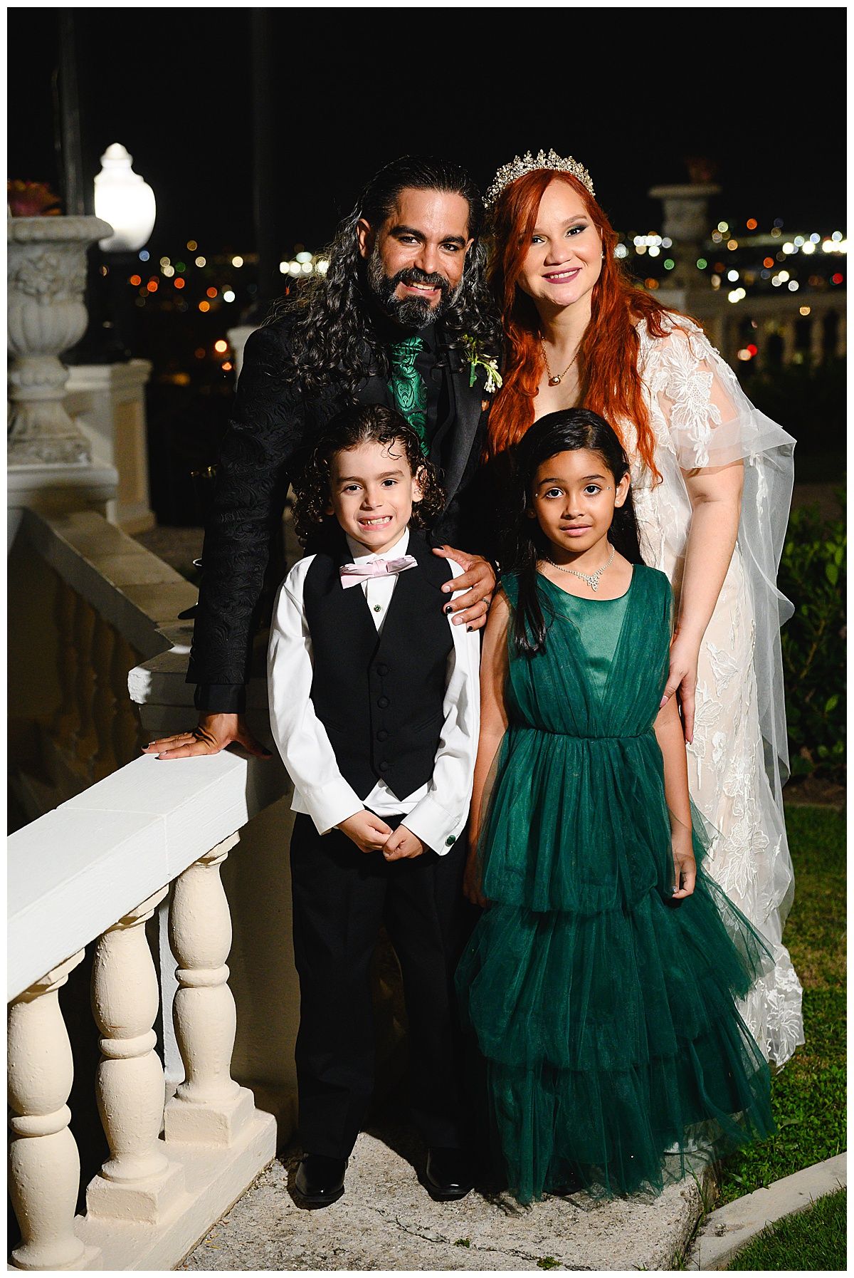 Wedding couple in formal attire pose with two children at night wedding, bride wearing tiara and groom in black suit.