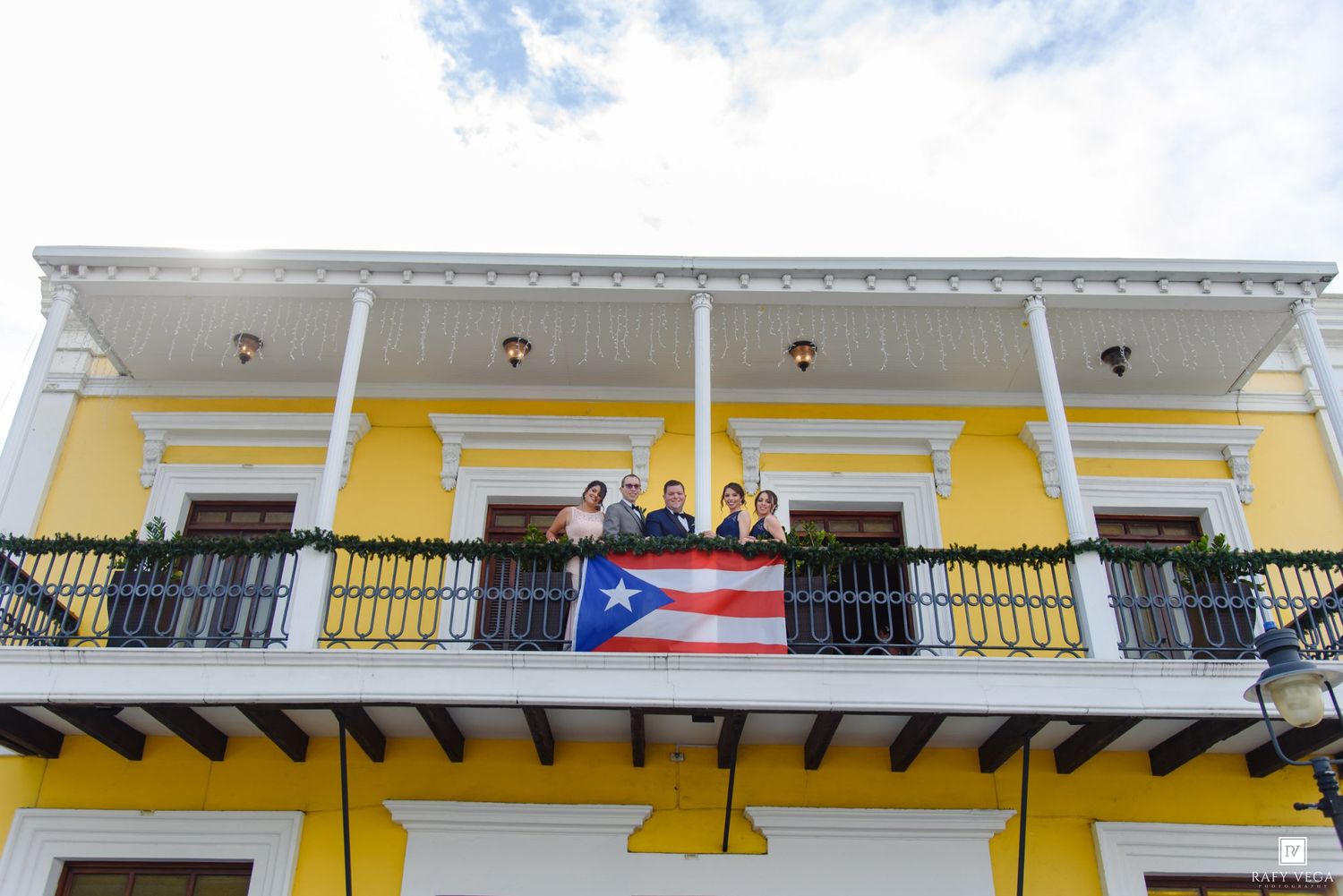 People on a yellow colonial building balcony displaying a Puerto Rico flag in Old San Juan.