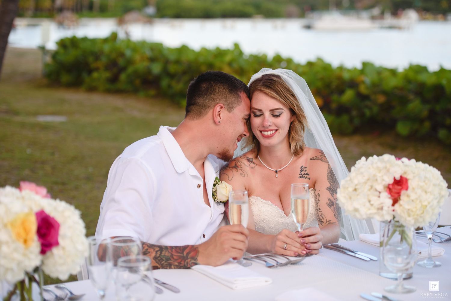 Bride and groom share a romantic moment at outdoor wedding reception table with champagne and floral arrangements.