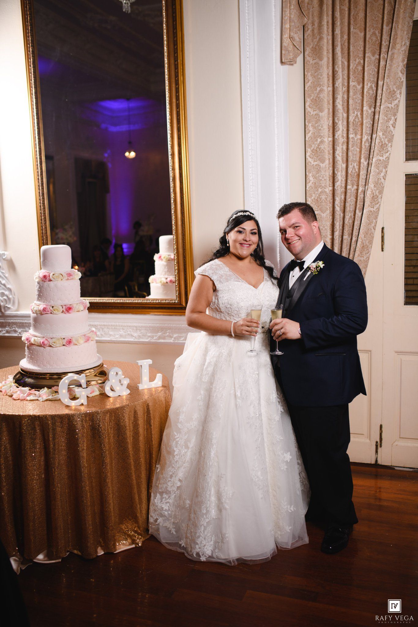 Bride and groom smiling beside a white wedding cake with G&L monogram letters at elegant reception venue.