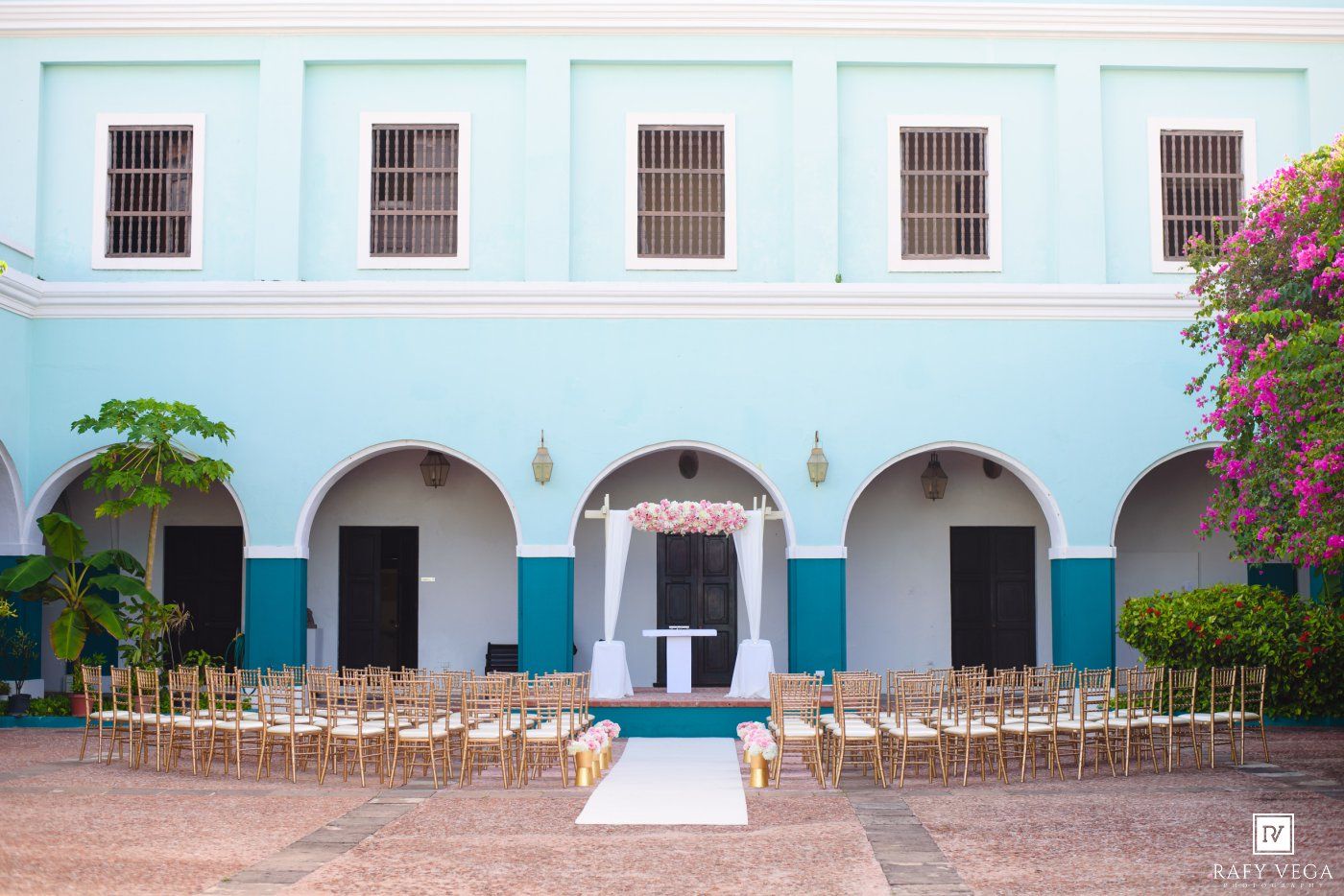 Outdoor wedding ceremony setup in a turquoise colonial courtyard with gold chairs and floral arch.