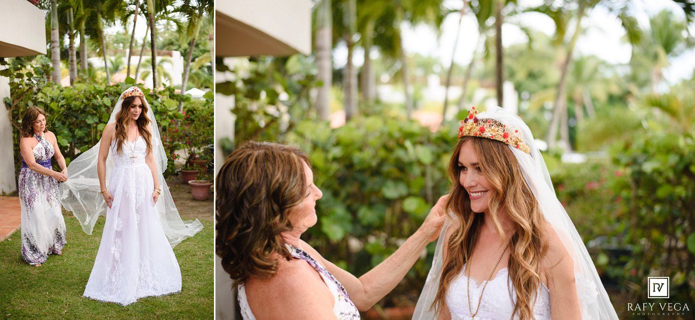 A bride in a white gown and veil stands outdoors while a woman adjusts her floral crown before the wedding ceremony.