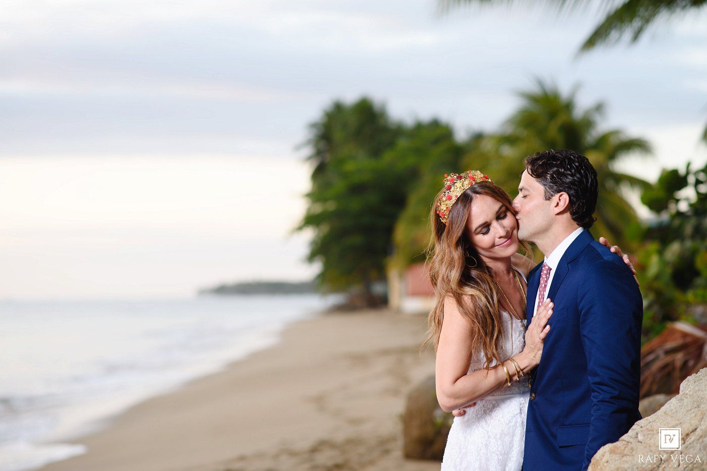 Couple embracing on tropical beach at sunset, woman in white dress with floral crown, man in navy suit, palm trees behind.
