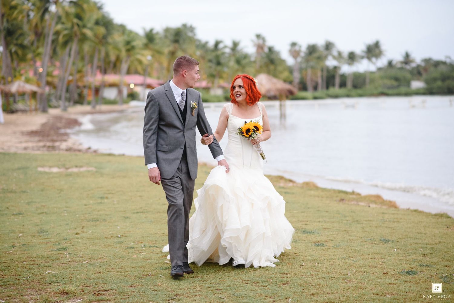 Bride with red hair holding sunflowers walks with groom in gray suit along tropical waterfront on their wedding day.