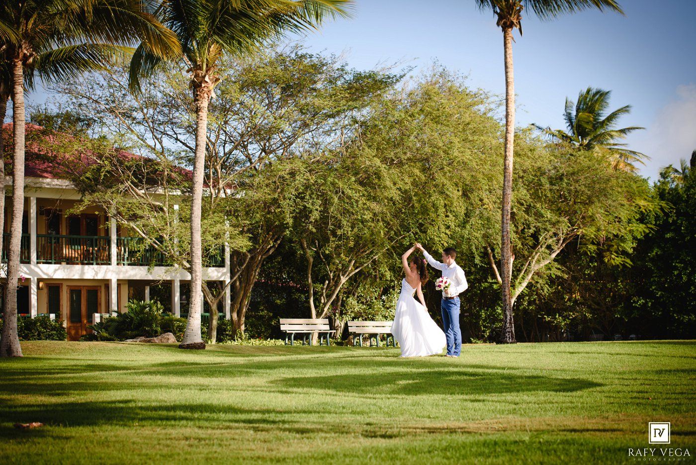 Bride and groom dance on lush green lawn surrounded by palm trees at tropical outdoor wedding venue.