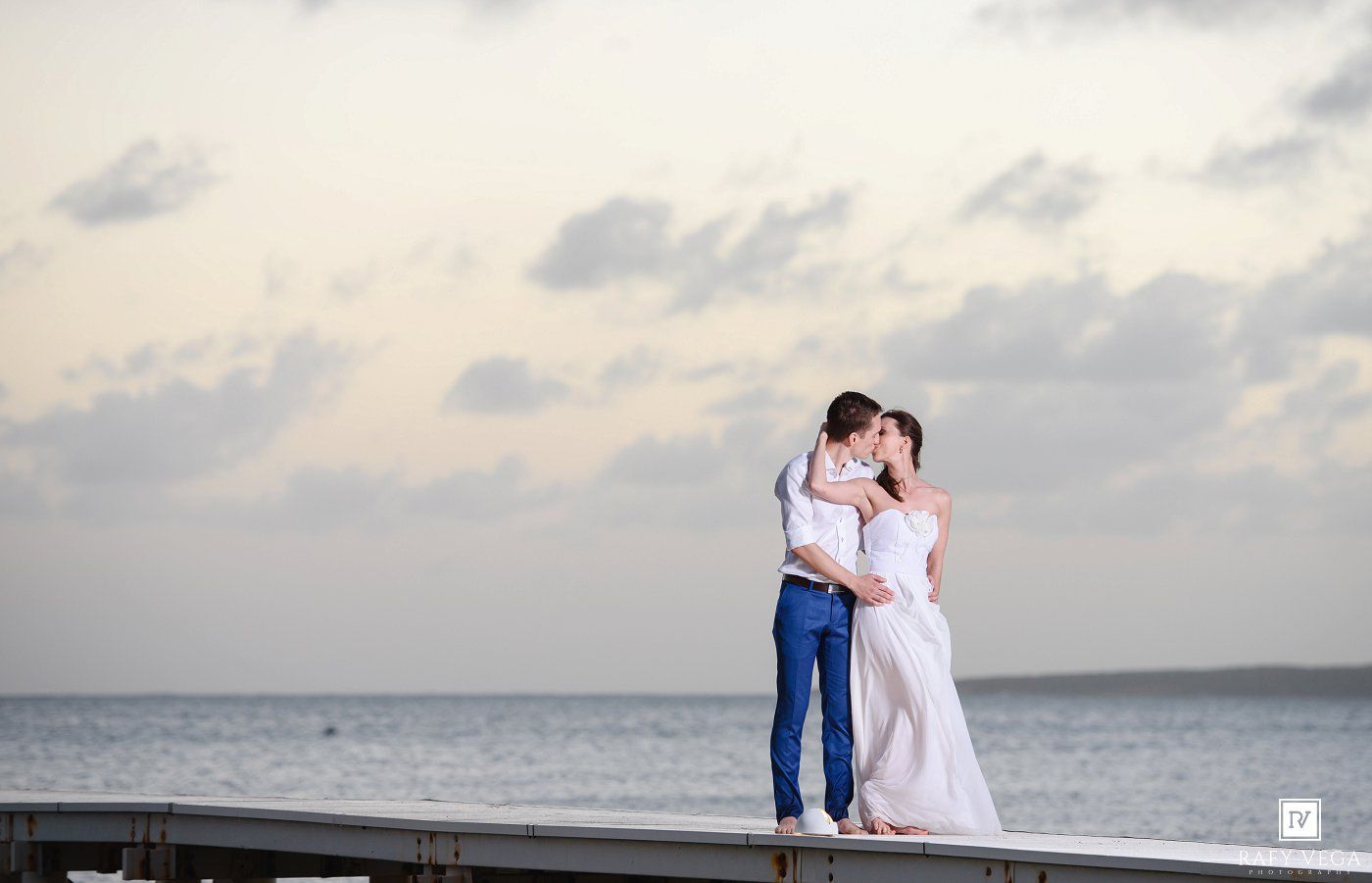 Couple sharing a romantic kiss on a seaside pier at sunset in wedding attire, with ocean and cloudy sky backdrop.