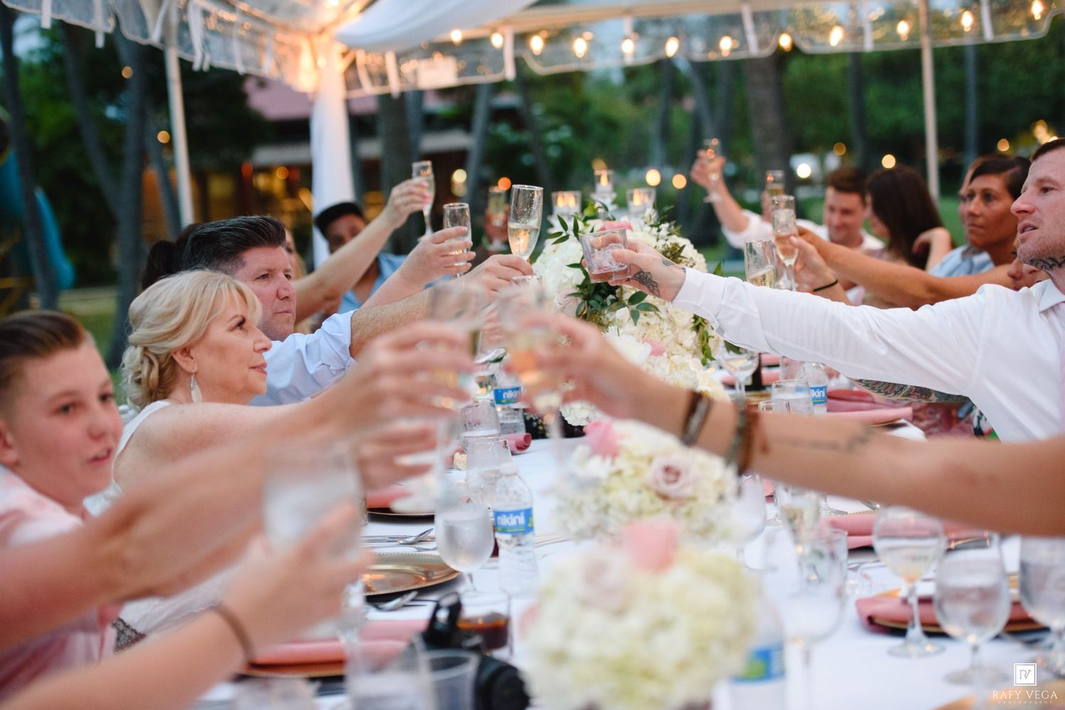 Guests raising champagne glasses in a toast at an elegant outdoor wedding reception dinner party.
