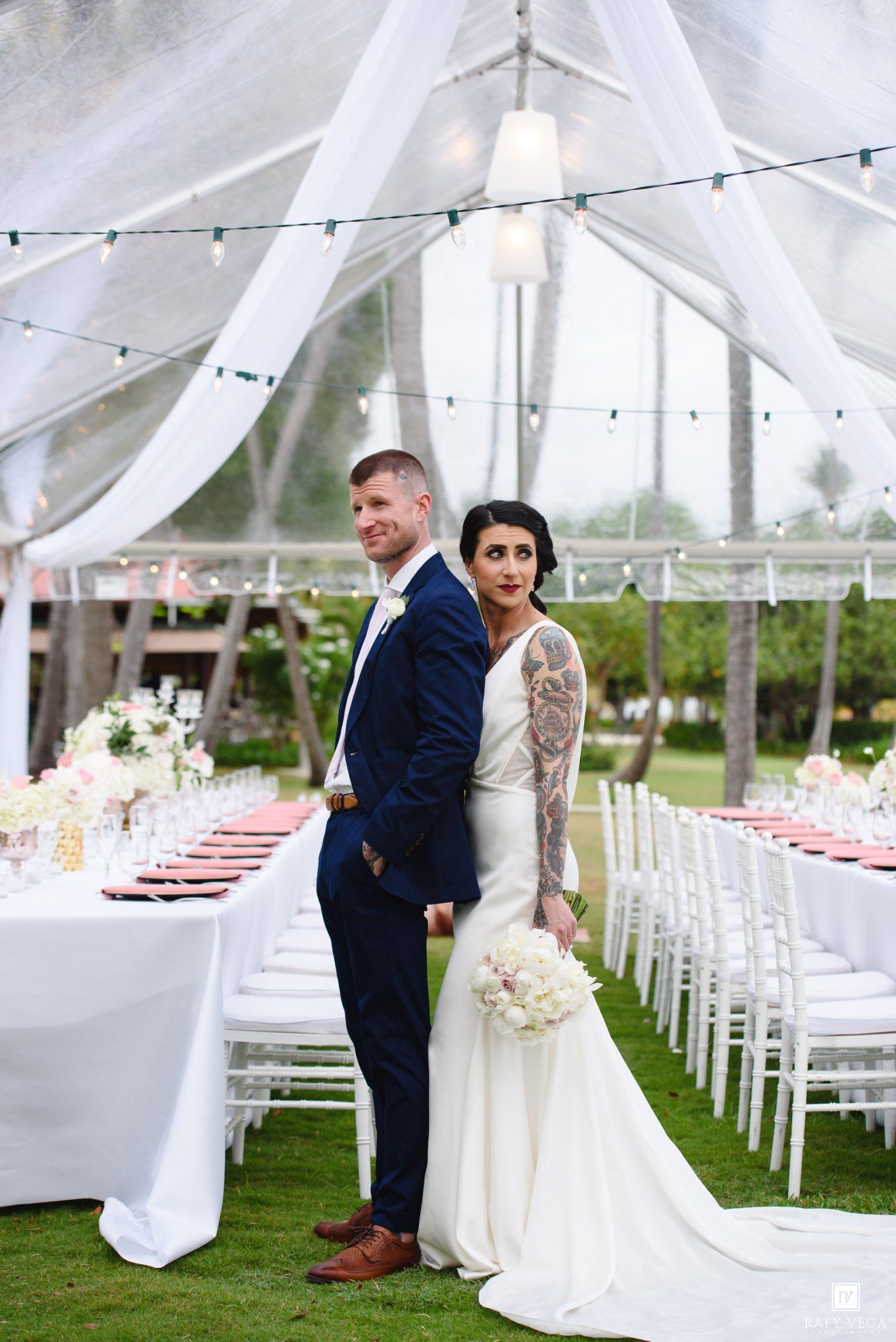 Bride in white gown with floral sleeve tattoos and groom in navy suit pose back-to-back under elegant draped wedding tent.