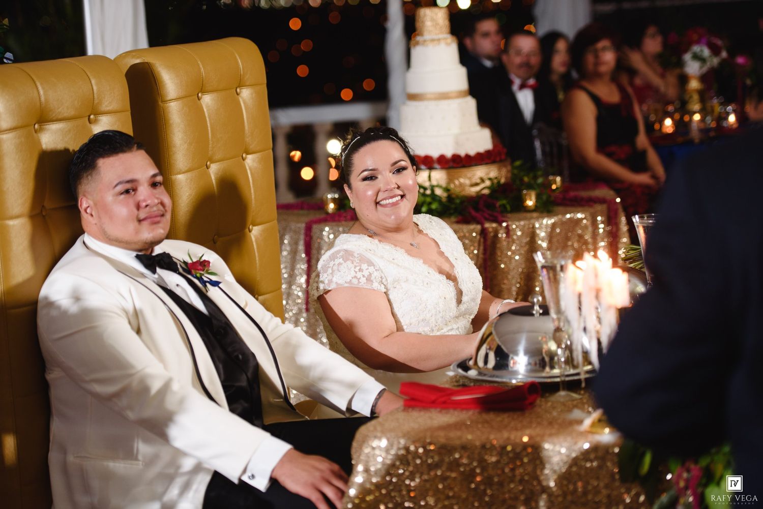 Bride and groom smiling at wedding reception seated at sweetheart table with tiered cake visible behind.