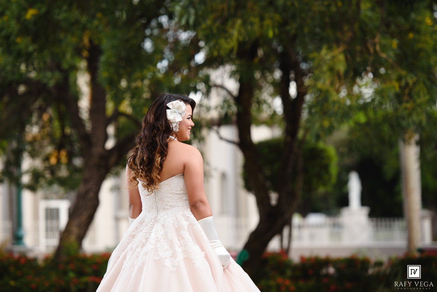 Bride in a strapless lace ball gown with floral hair accessory poses outdoors near trees and a white building.