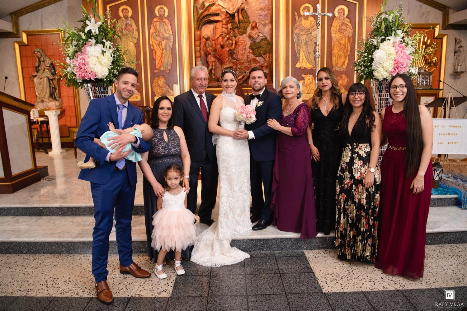 Wedding party poses in church with bride in white gown, groom in blue suit, bridesmaids in burgundy dresses, and family members.