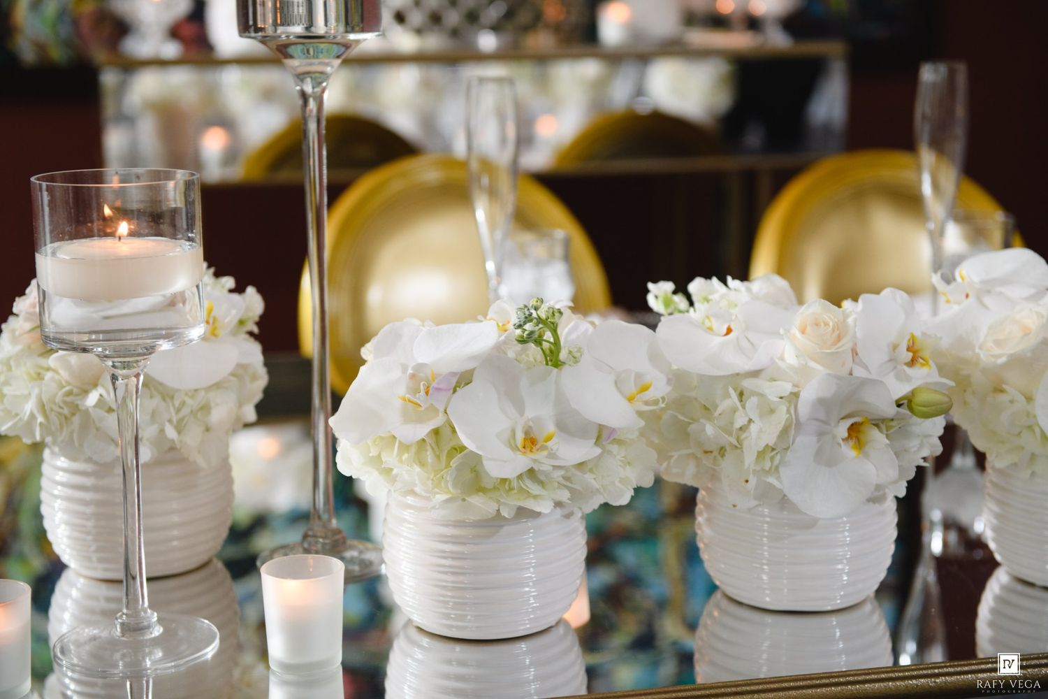 Elegant white floral centerpieces with orchids and hydrangeas in white vases on a mirrored table with candles.